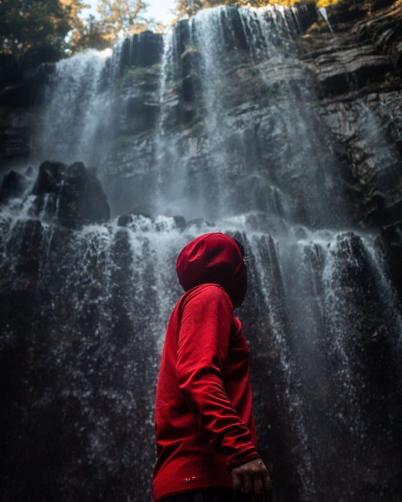 A man in a red hoodie stands in front of a waterfall. - Waterfall