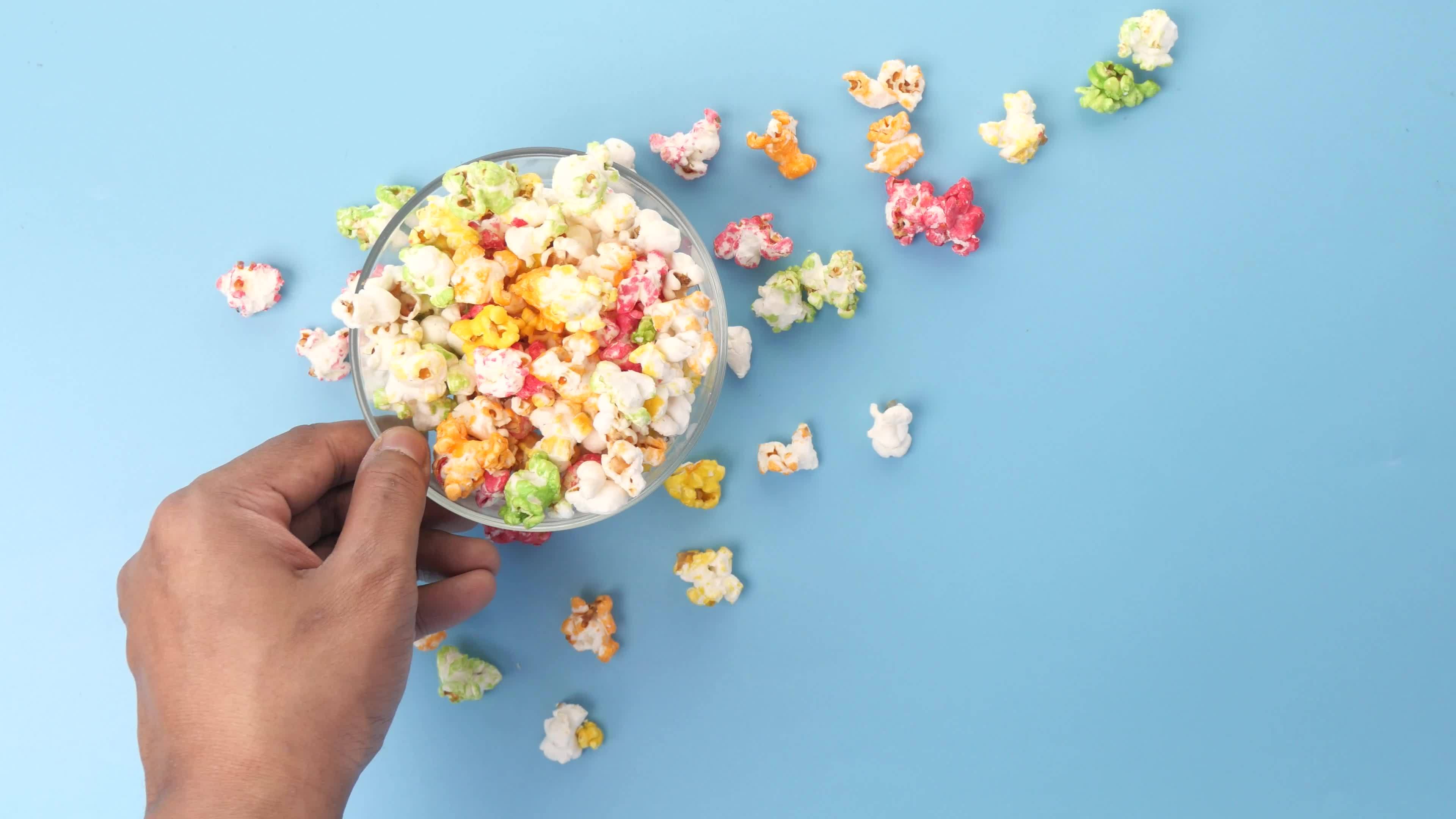 A hand holding a bowl of popcorn with some popcorn scattered around - Popcorn