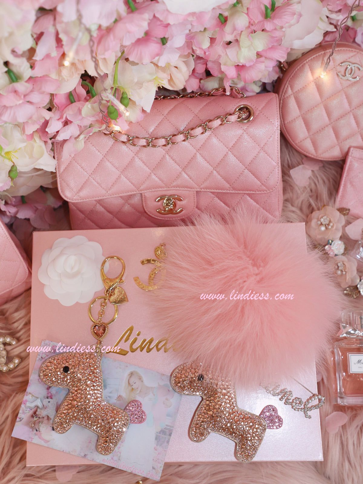 A pink Chanel bag, a pink fluffy keychain, and a pink card holder are laid out on a fur blanket. - Bling