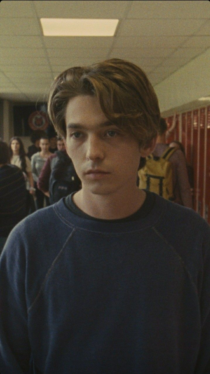 A young man with brown hair and a blue sweater stands in a school hallway - Austin Abrams