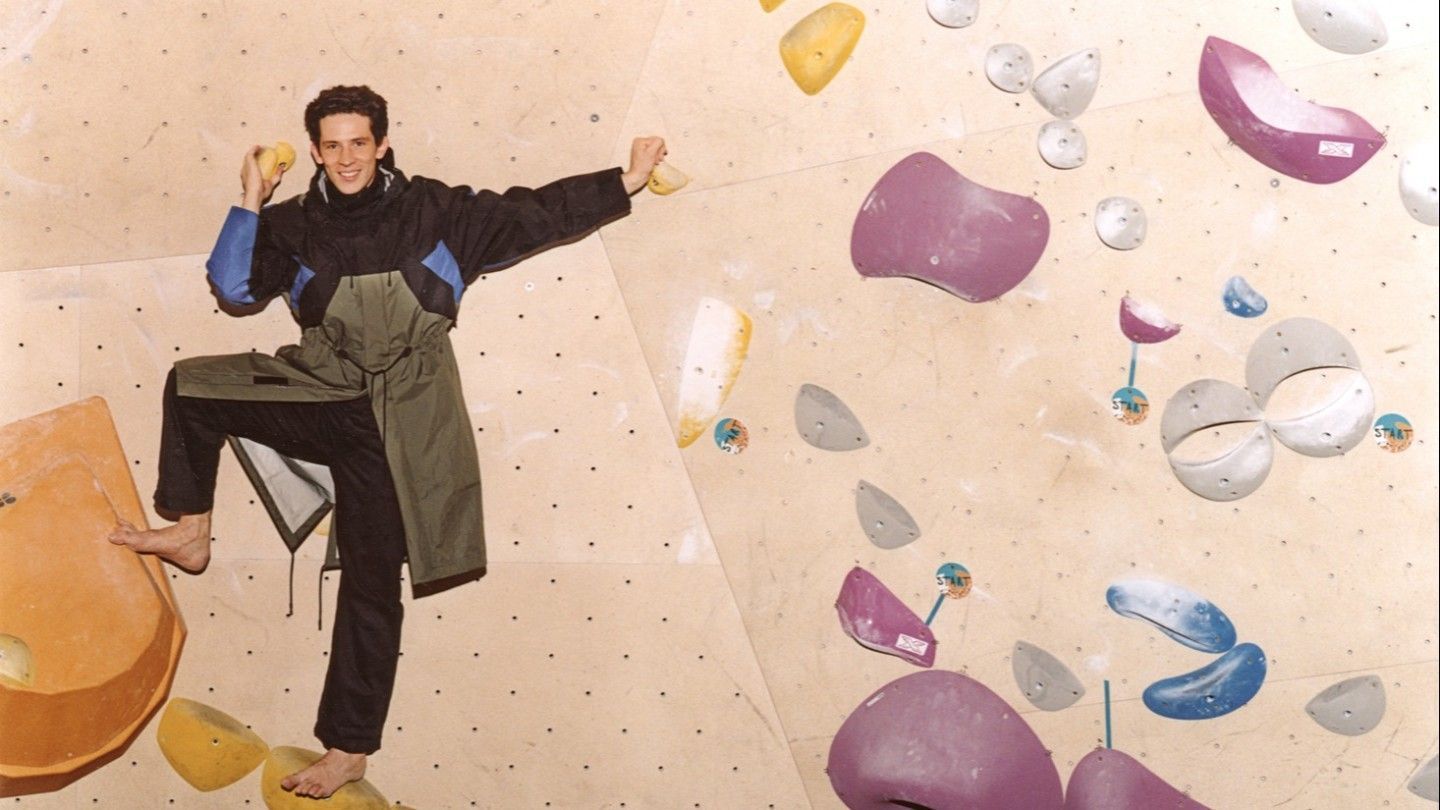 A young man in a black raincoat stands on a rock climbing wall - Josh O'Connor