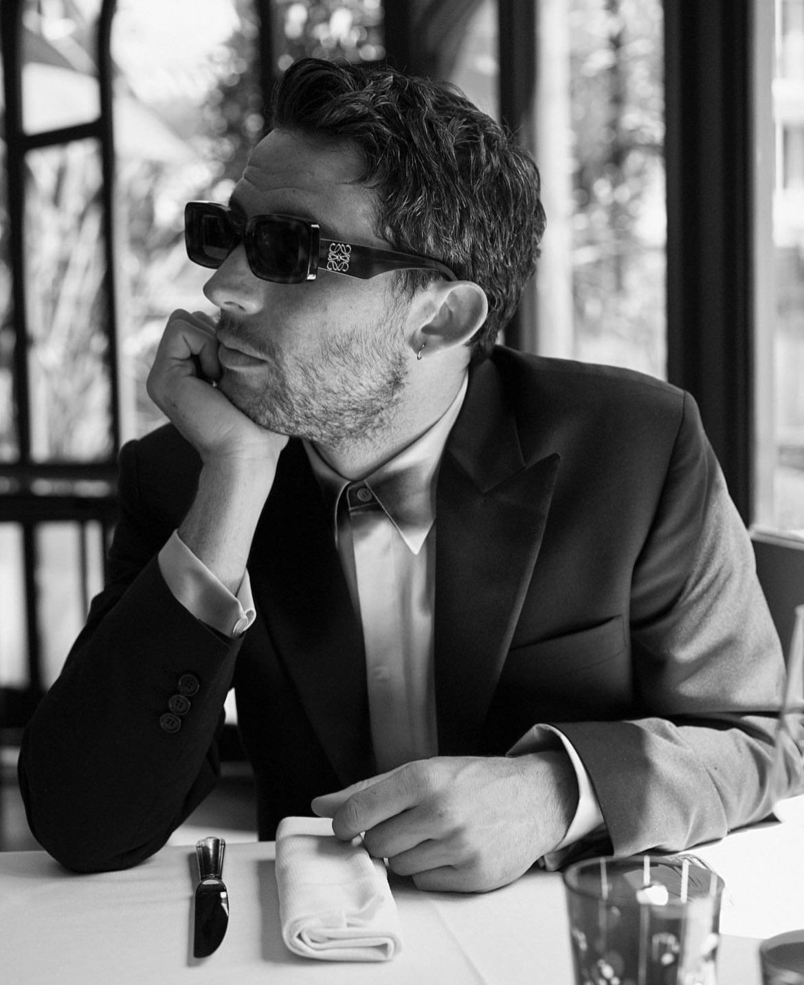 A man in a suit and sunglasses sits at a table. - Josh O'Connor
