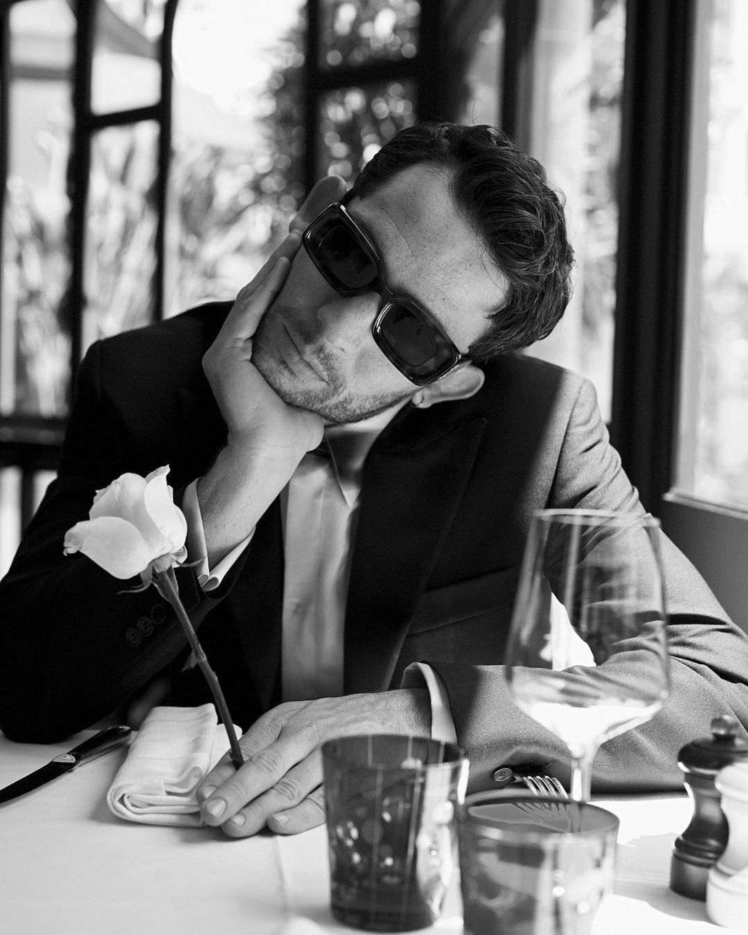 A man in a suit and sunglasses sits at a table with a flower in a wine glass. - Josh O'Connor