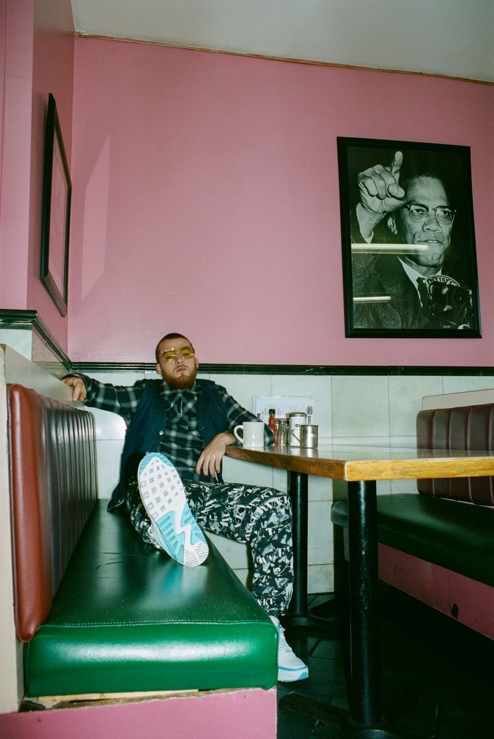 A man sitting on a booth in a diner with a picture of Malcolm X on the wall behind him - Angus Cloud