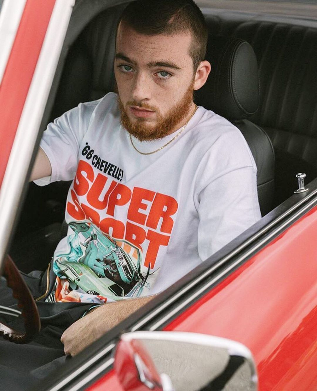 Mac Miller wearing a white tee with a graphic print on the front sitting in a car. - Angus Cloud