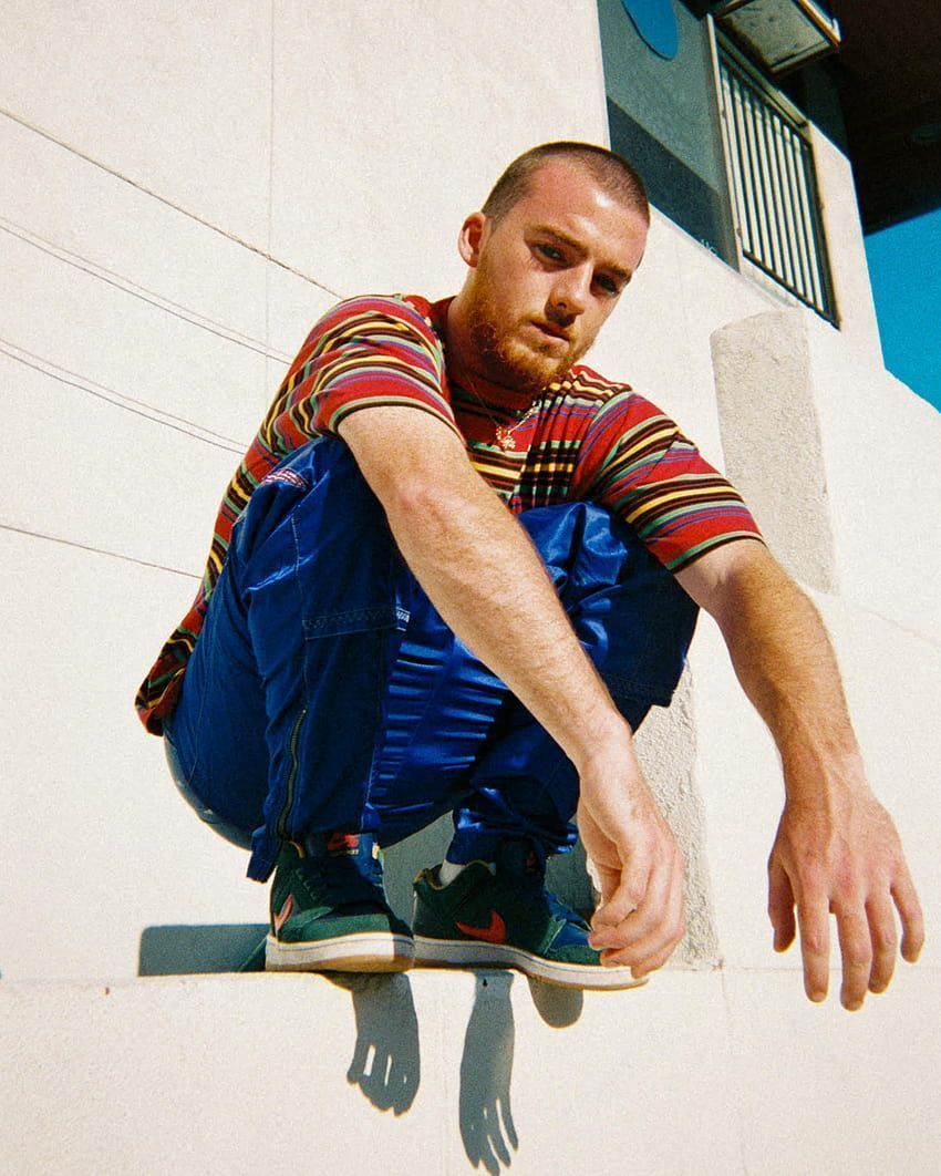 A man with a beard in a red striped shirt and blue pants is on a skateboard. - Angus Cloud