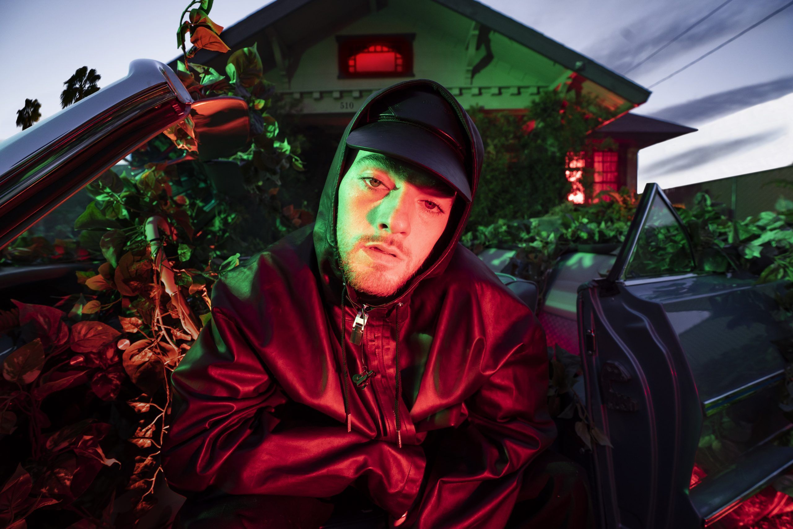 Mac Miller in a red jacket and black hat sitting in the passenger seat of a car - Angus Cloud