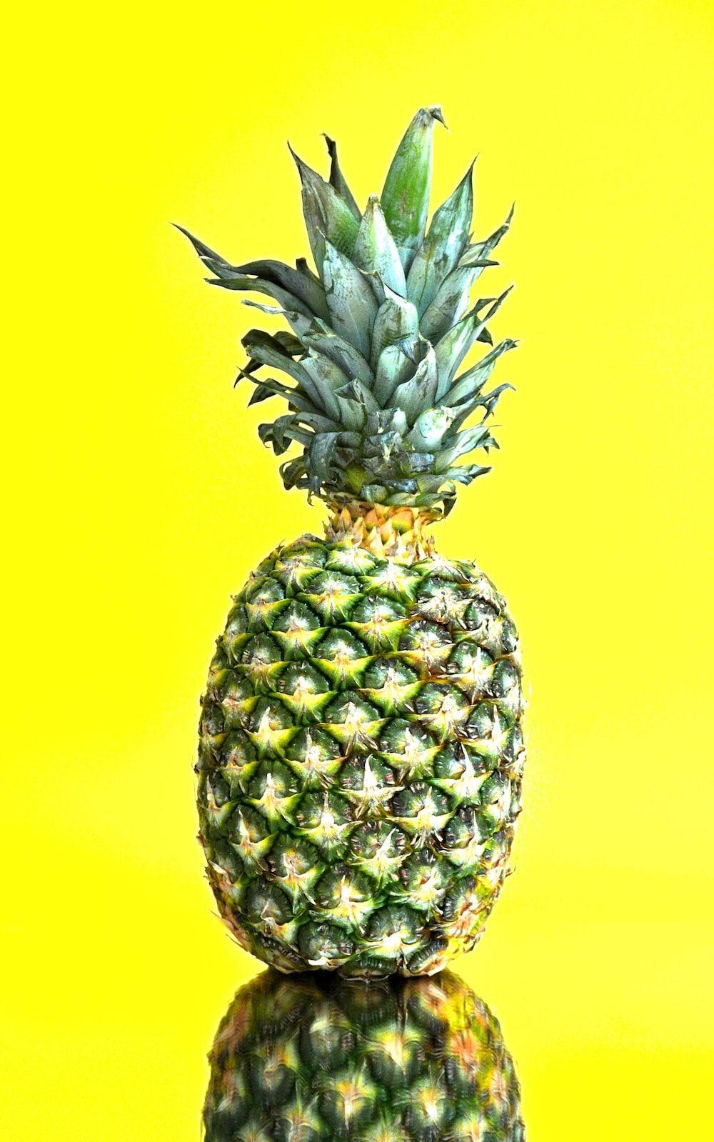 A pineapple with a reflection stands in front of a yellow background. - Pineapple