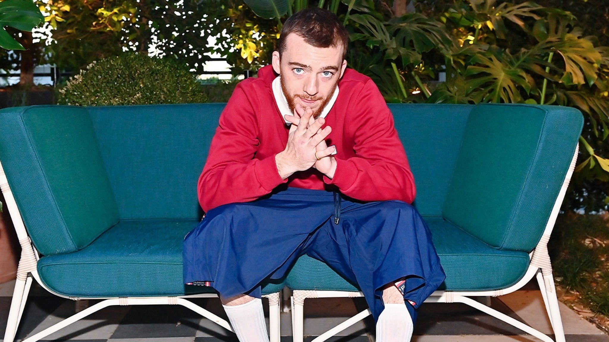 Sam Smith in a red sweater and blue pants sitting on a blue couch - Angus Cloud