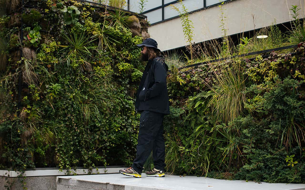 A man in black jacket and hat standing on concrete steps in front of a wall covered in plants. - Gorpcore
