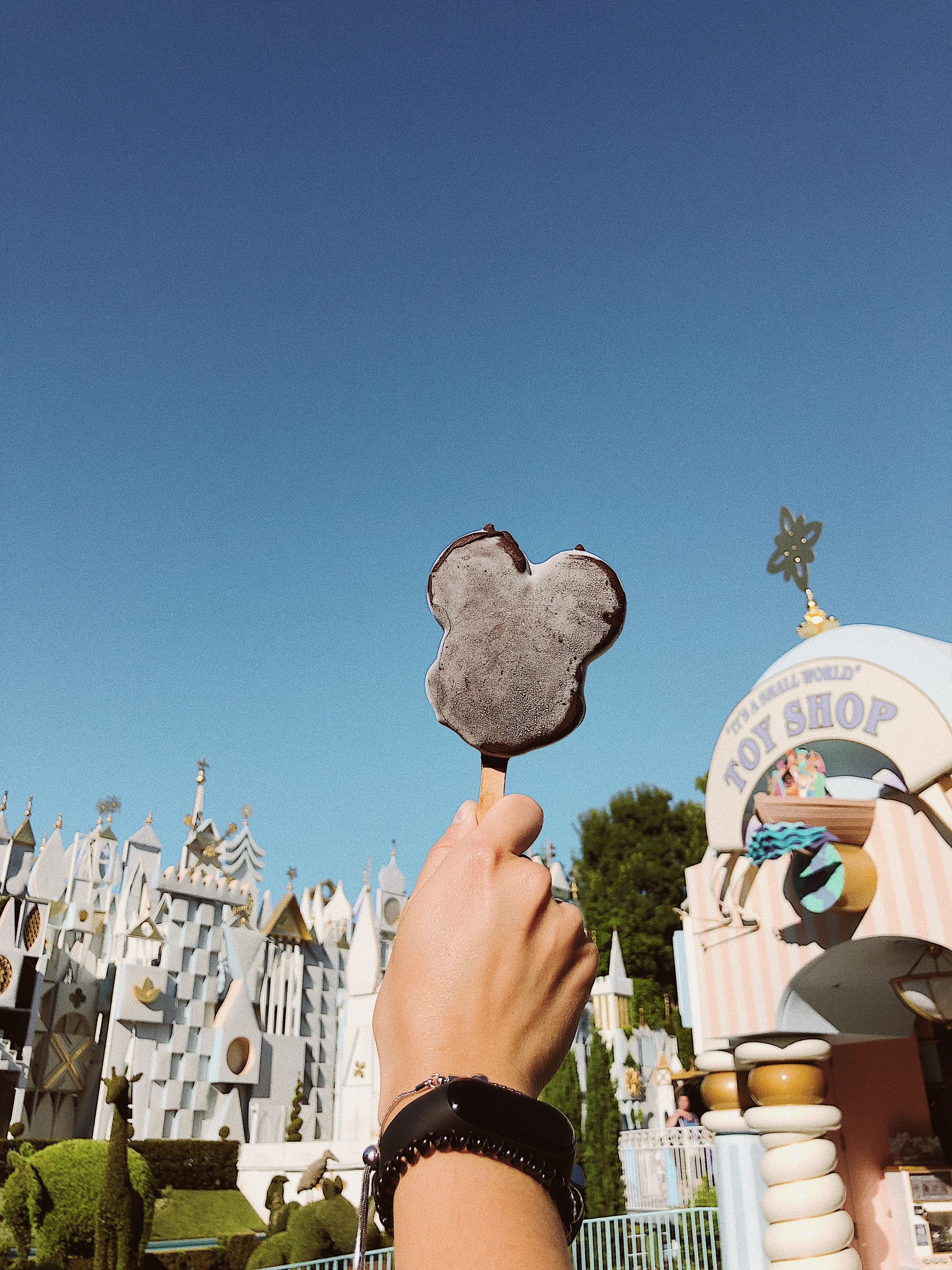 A hand holding a Mickey Mouse shaped chocolate ice cream bar in front of a castle. - Disneyland