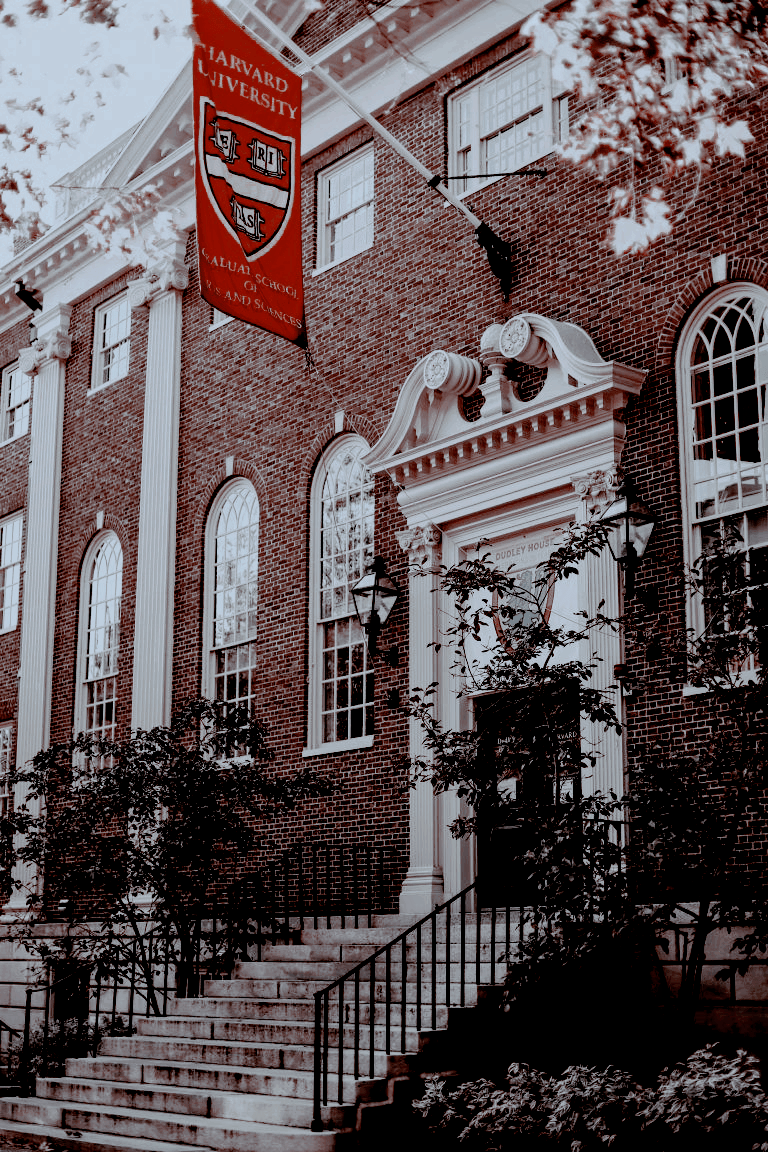 A red and white Harvard University banner hangs from a brick building. - Harvard