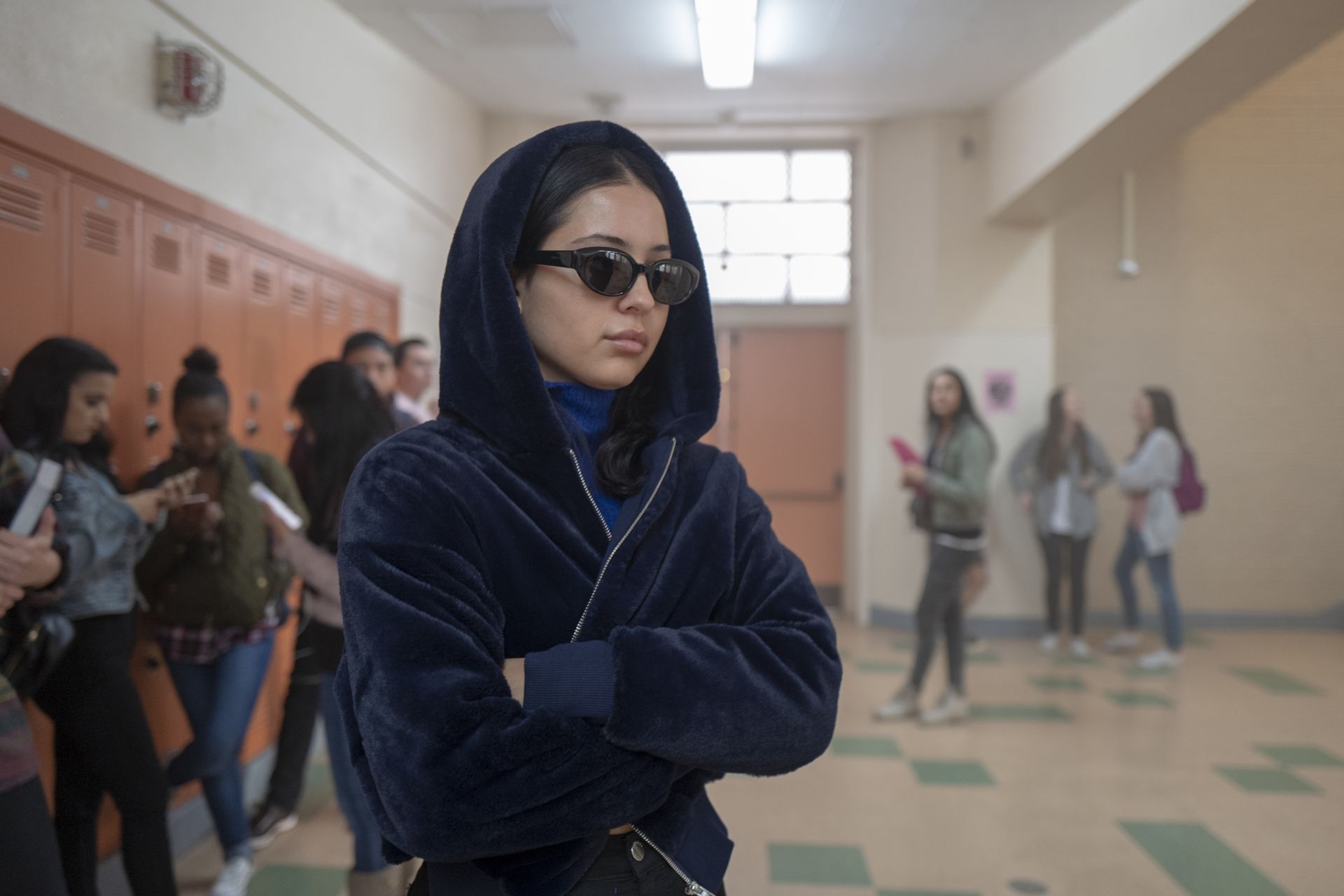 A young Asian woman with black sunglasses and a blue hoodie stands in a school hallway with her arms crossed. - Euphoria