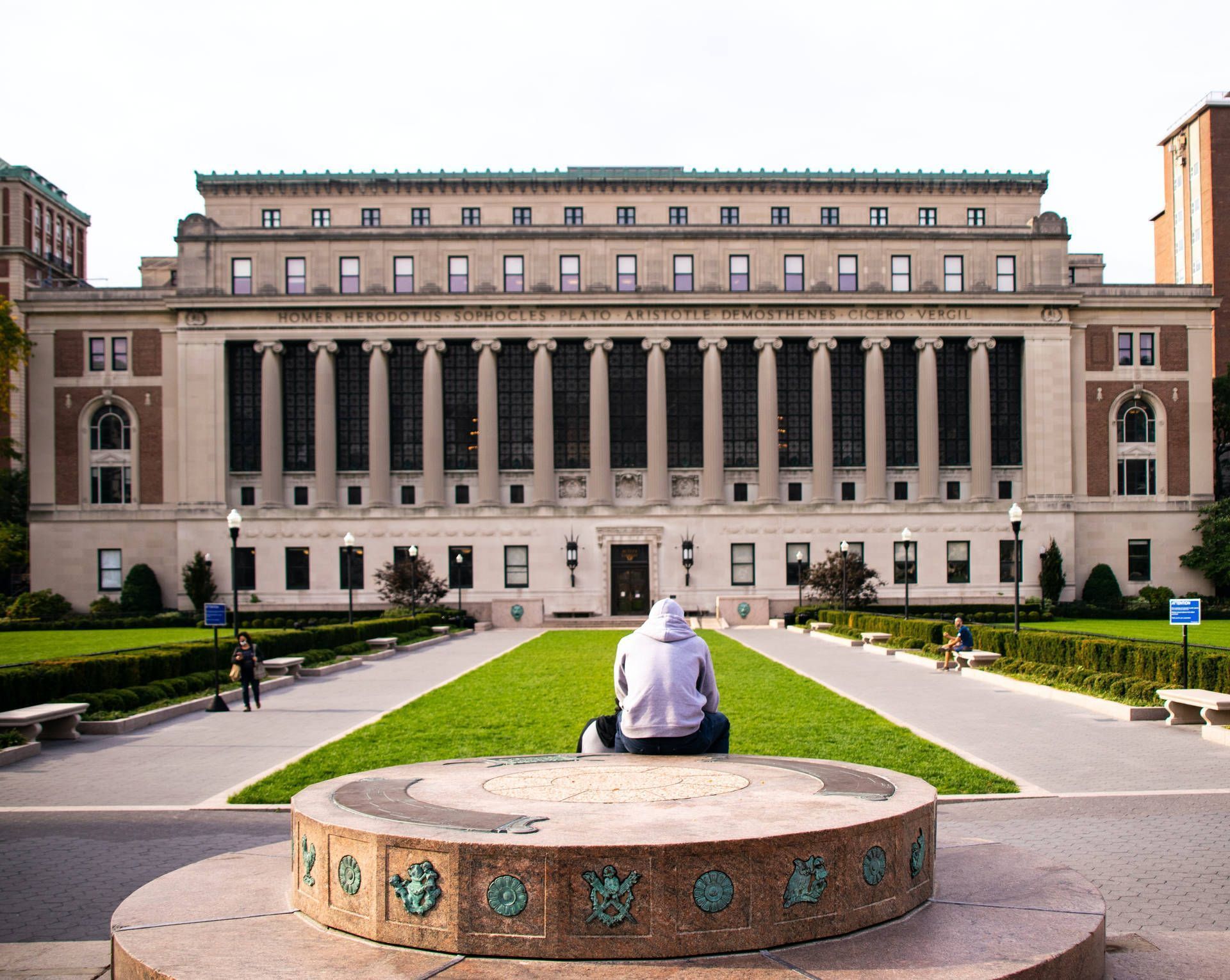 A student sits on a bench in front of a large building at Columbia University. - Columbia University