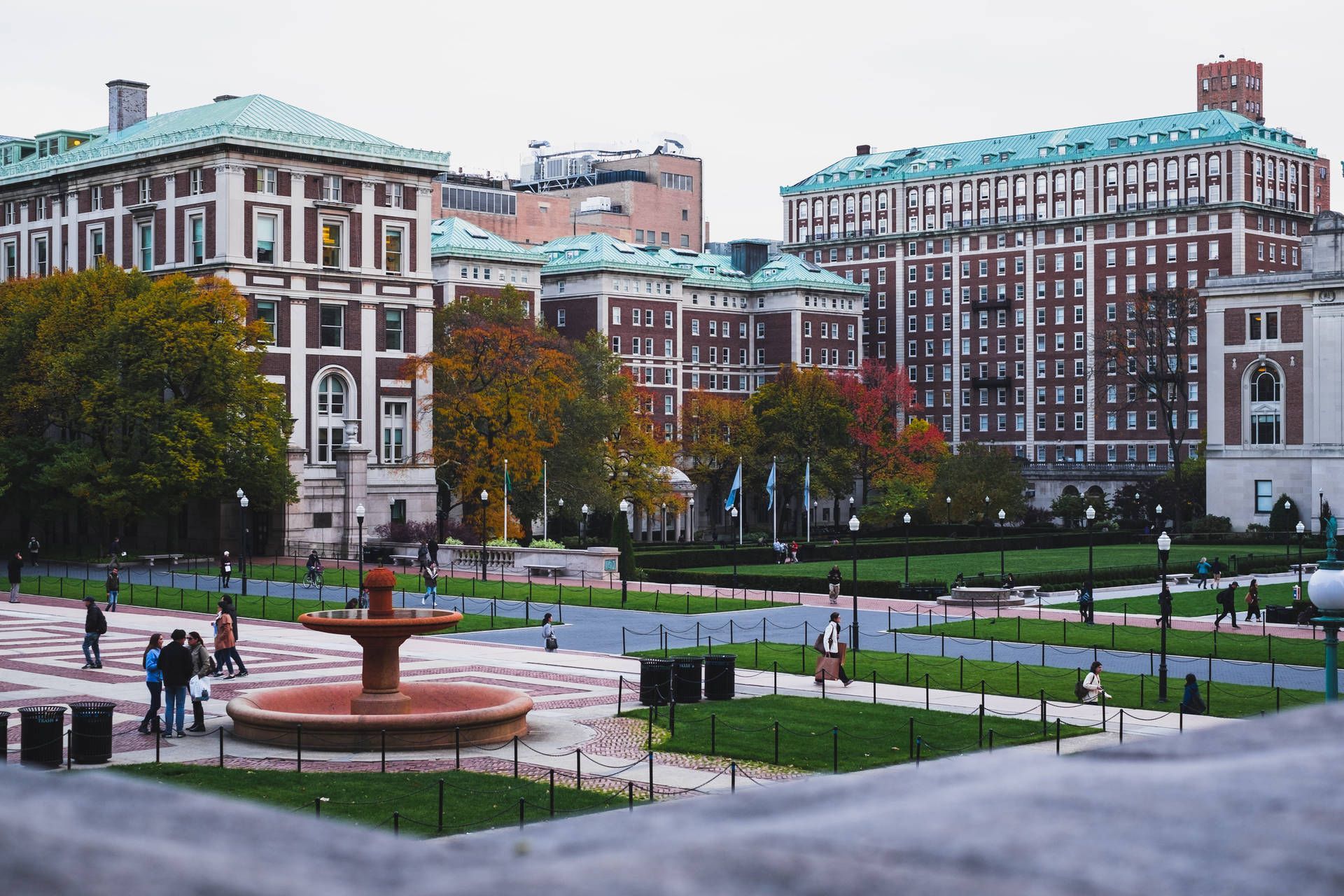 A red fountain in the middle of a green lawn in front of a large building. - Columbia University