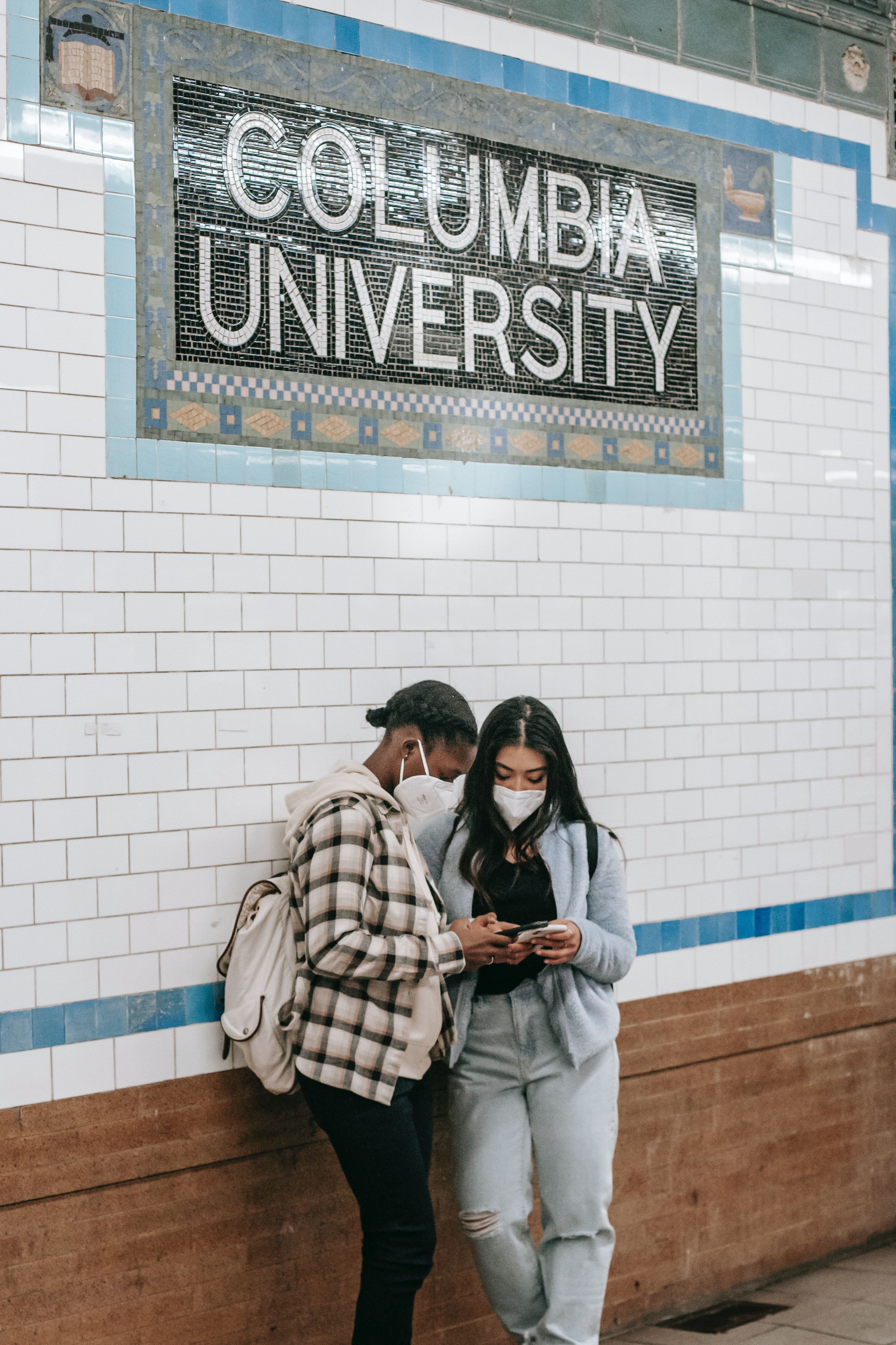 Two women wearing masks stand in front of a Columbia University sign in a subway station. - Columbia University