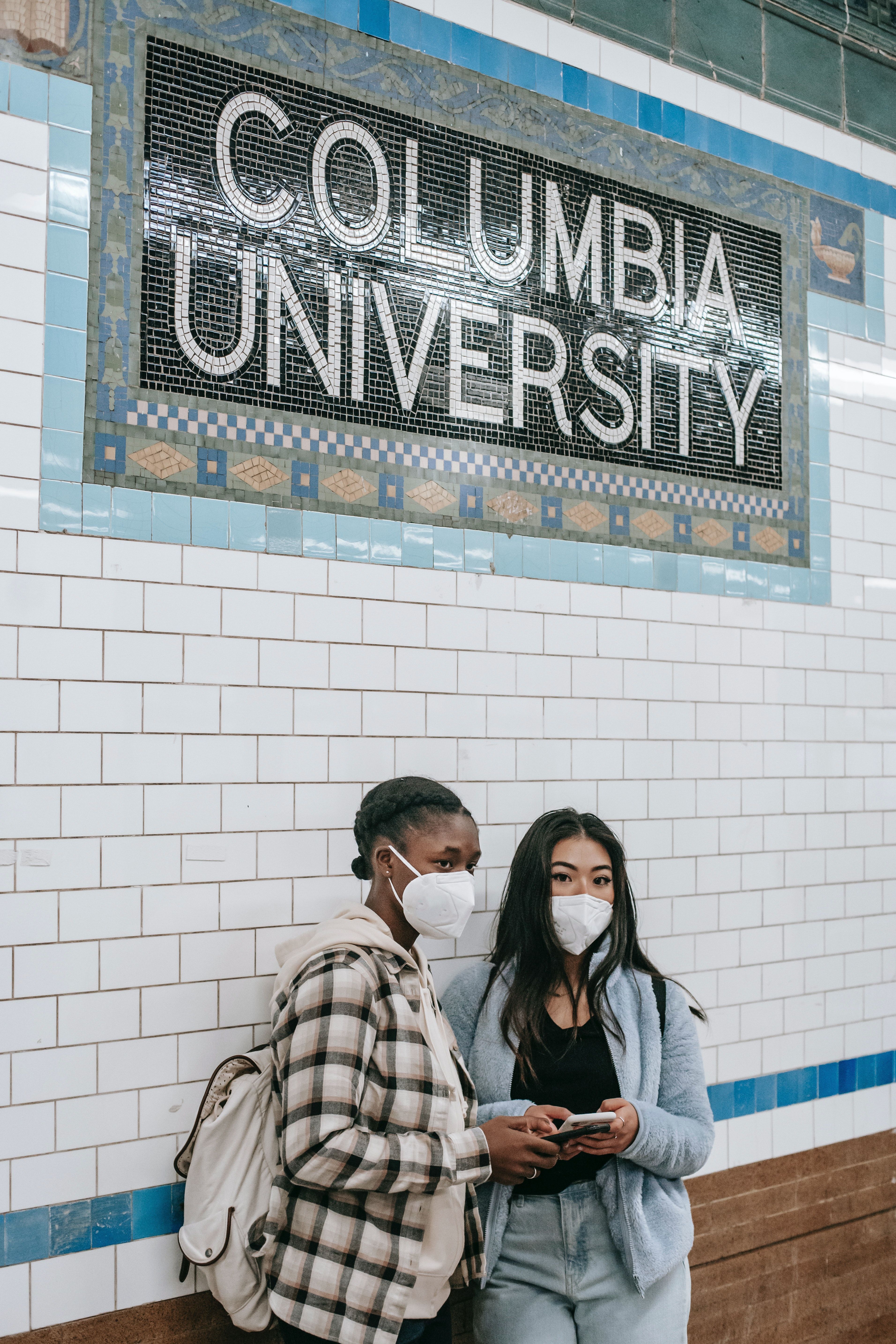 Two women wearing masks stand under a Columbia University sign in a subway station. - Columbia University