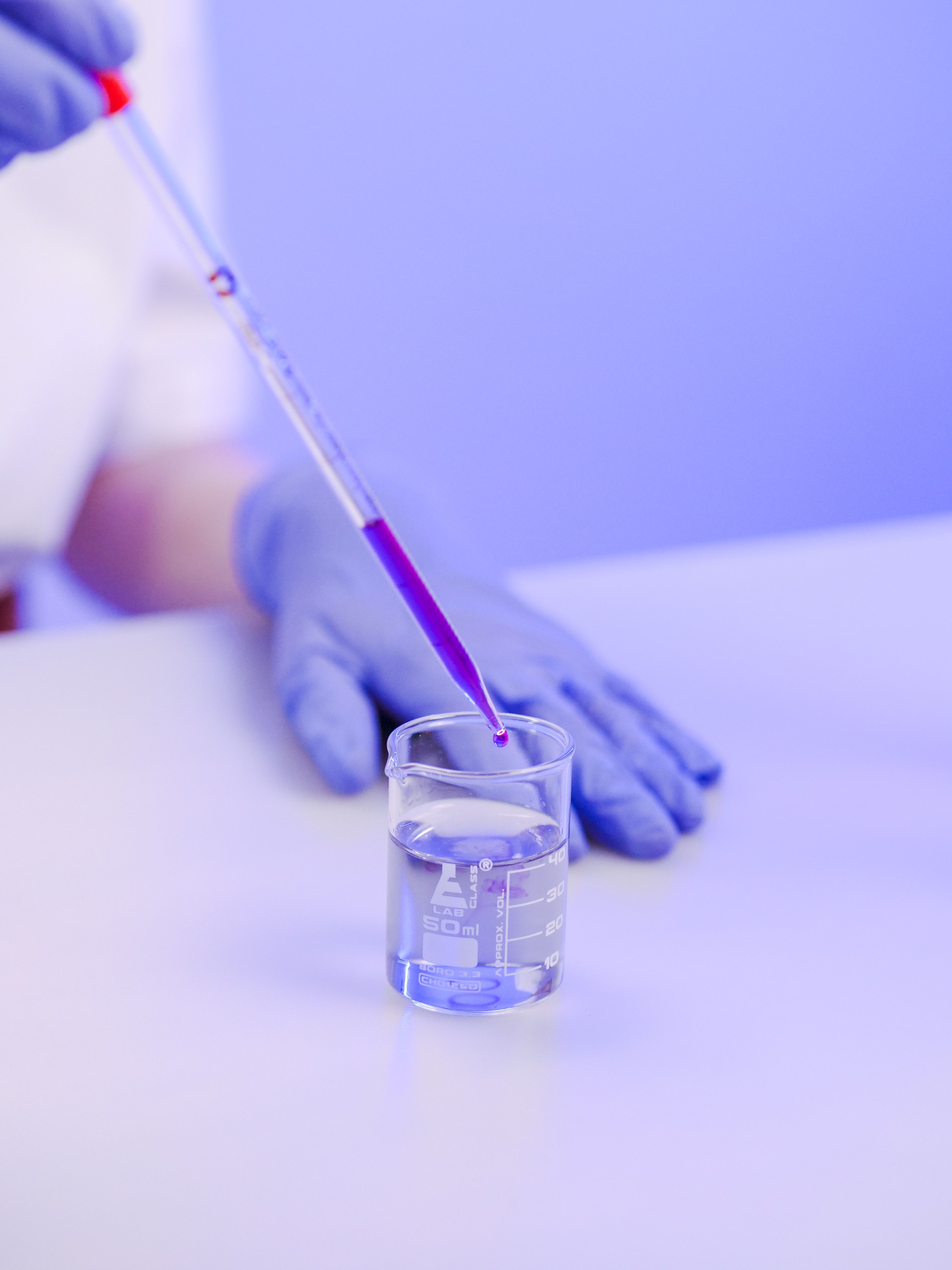 A person wearing blue gloves and holding a dropper with purple liquid. - Chemistry