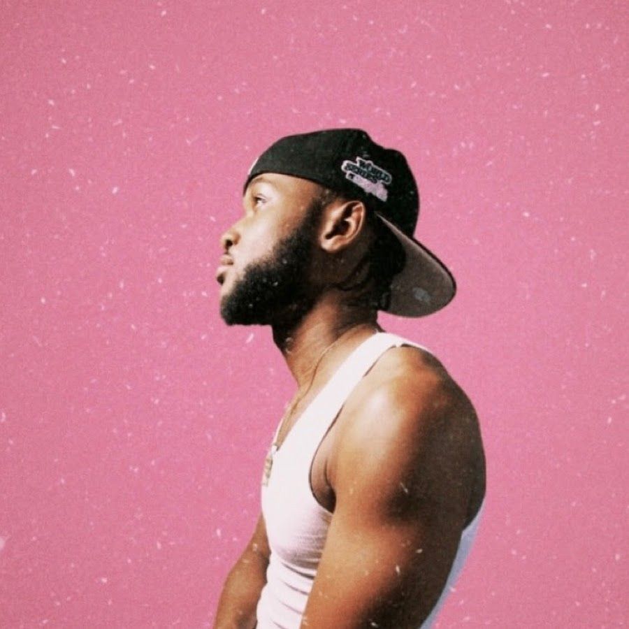A man with a beard and a baseball cap looking to the side against a pink background - Chance the Rapper