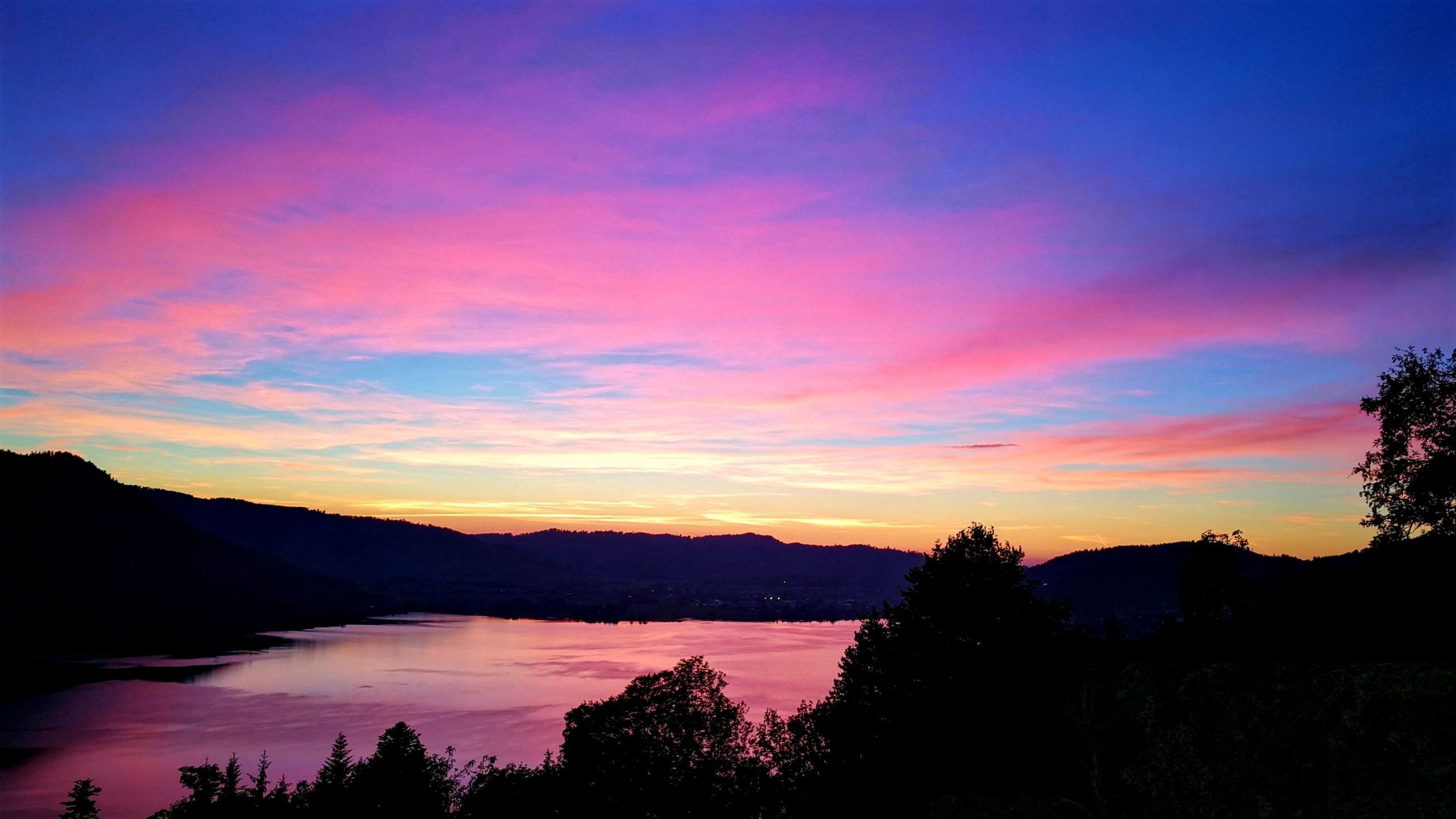 The sun sets over a lake, casting a pink and blue glow over the sky. - 2560x1440