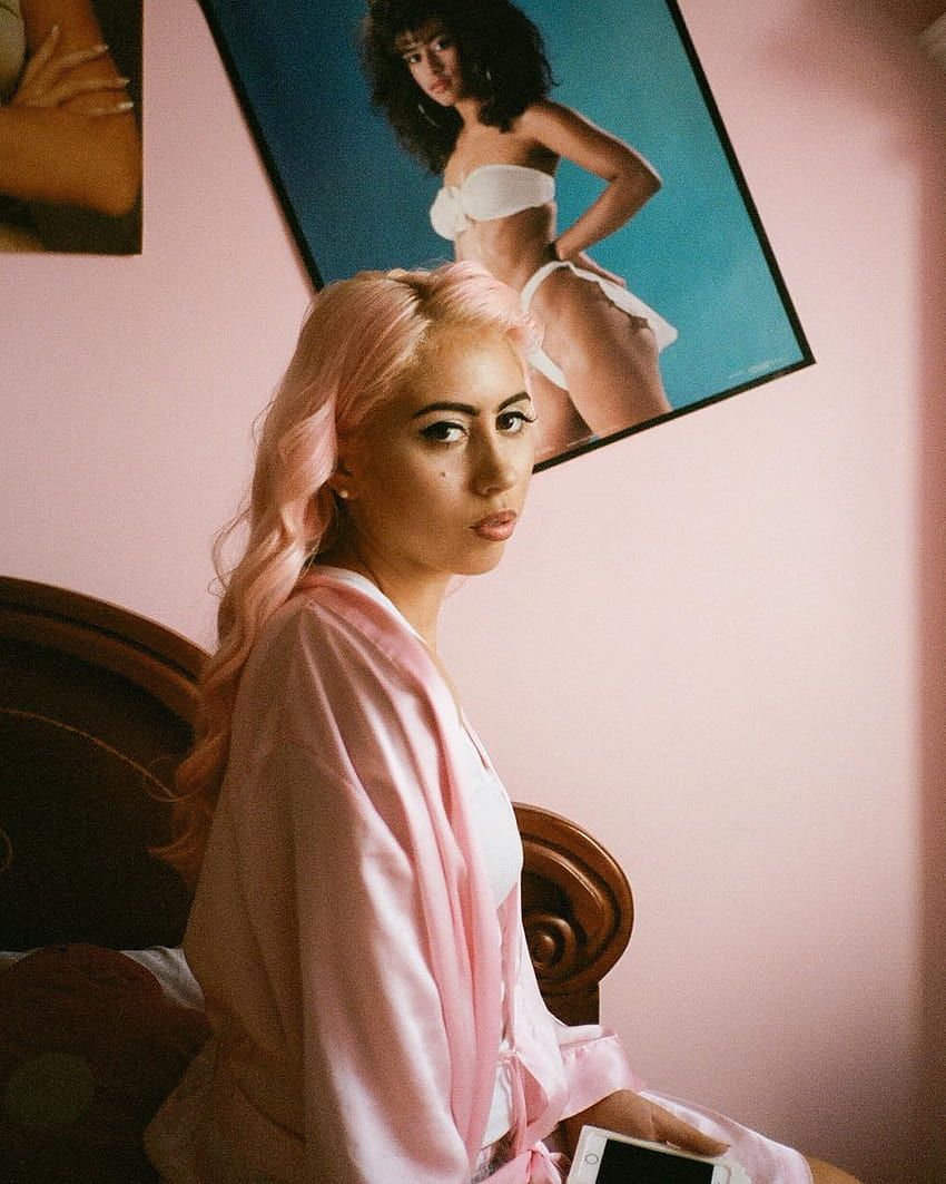 A woman with pink hair sitting on a bed - Kali Uchis