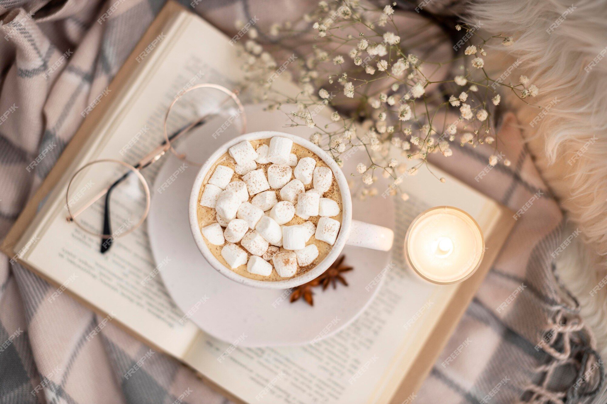 A cup of hot chocolate with marshmallows on top, a book, a candle and glasses on a blanket. - Marshmallows