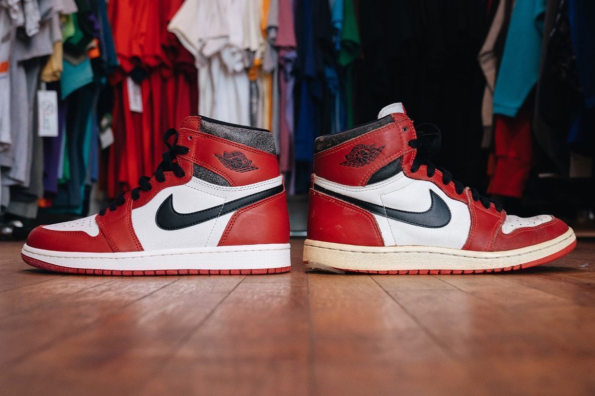 A pair of red and white Nike Air Jordan 1 shoes sit on a wooden floor in front of a rack of hanging clothes. - Michael Jordan, Air Jordan 1, Air Jordan 5