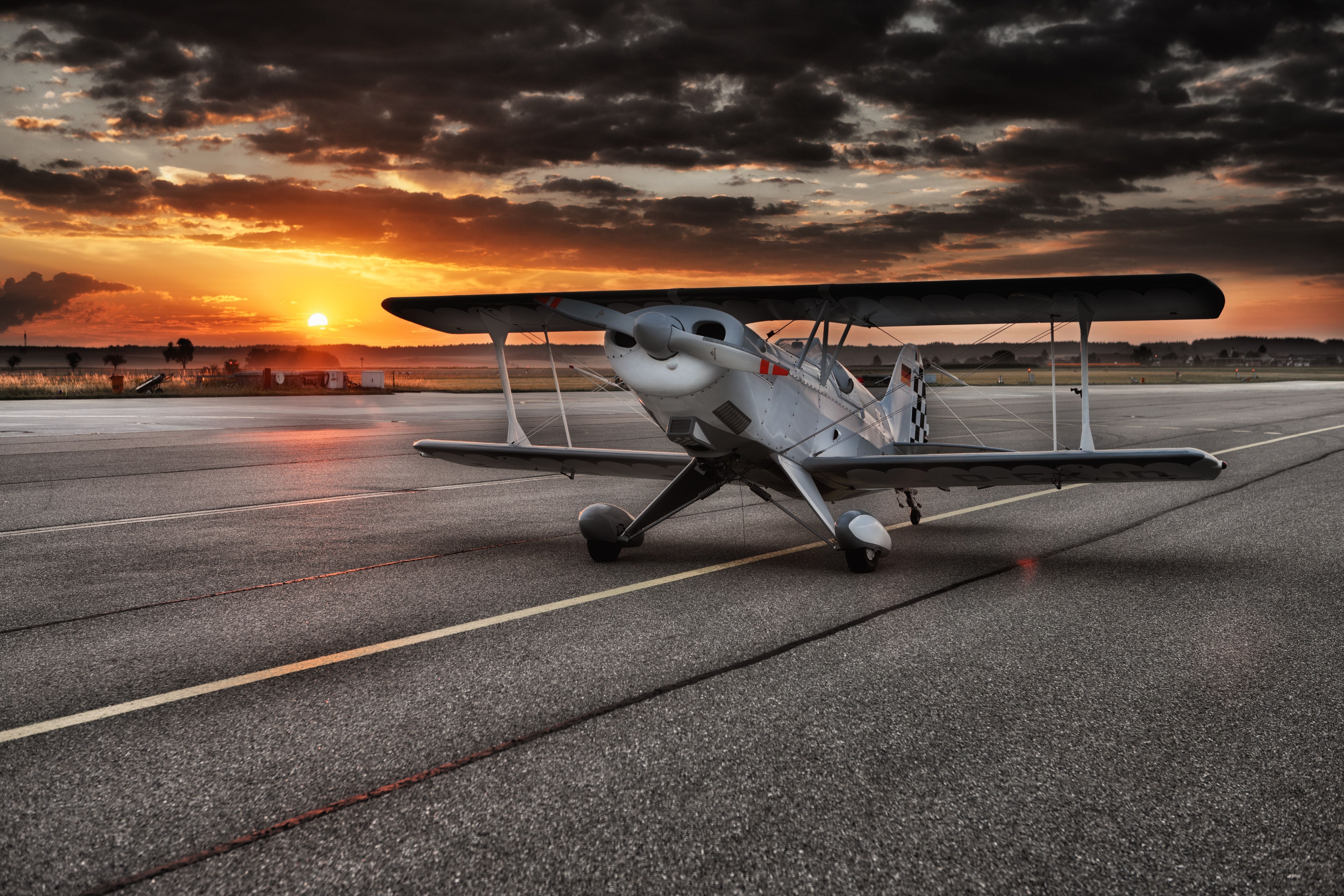 A biplane sits on the runway as the sun sets behind it. - Airplane