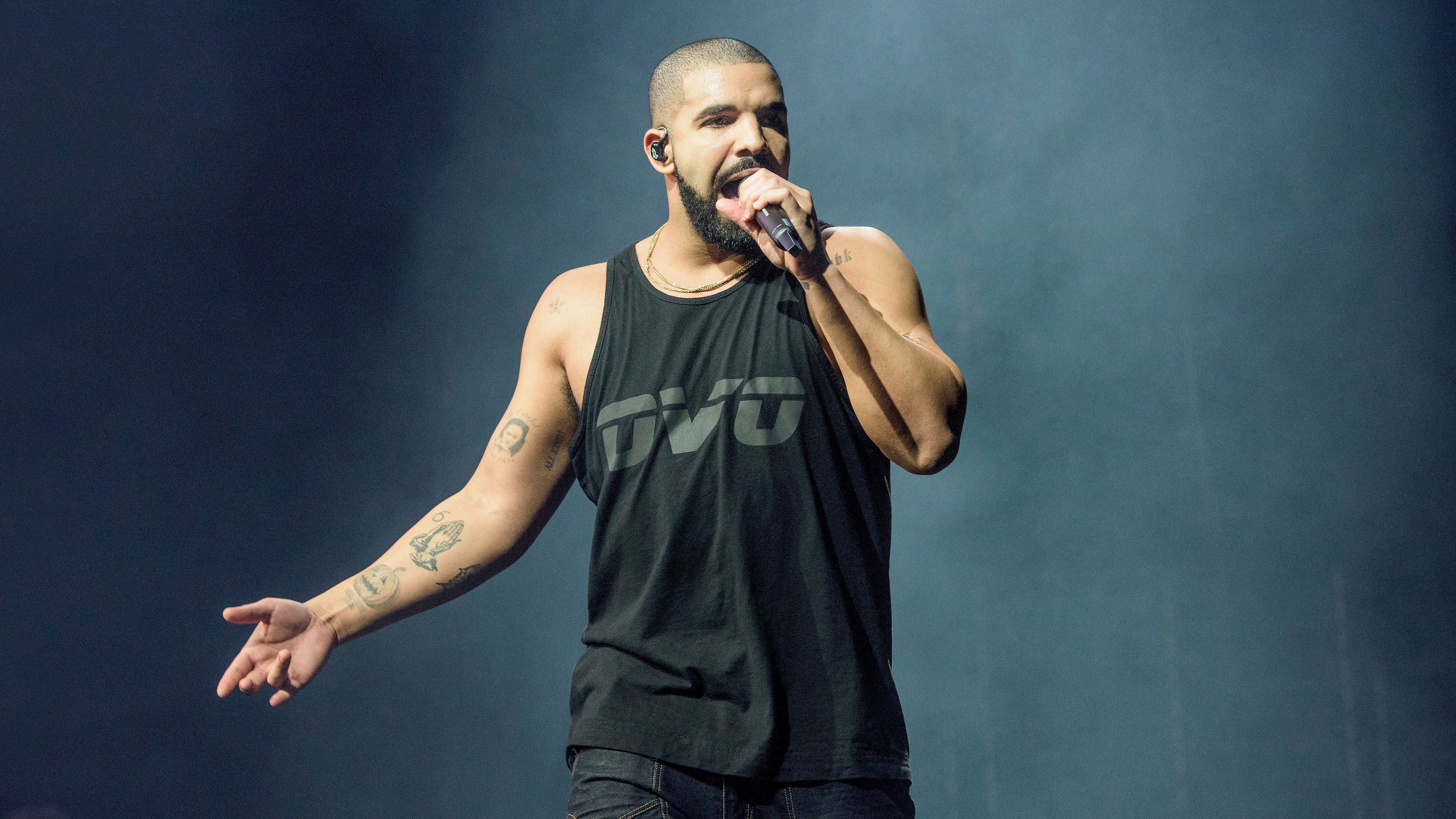 A man in black shirt and tank top is holding up his microphone - Drake