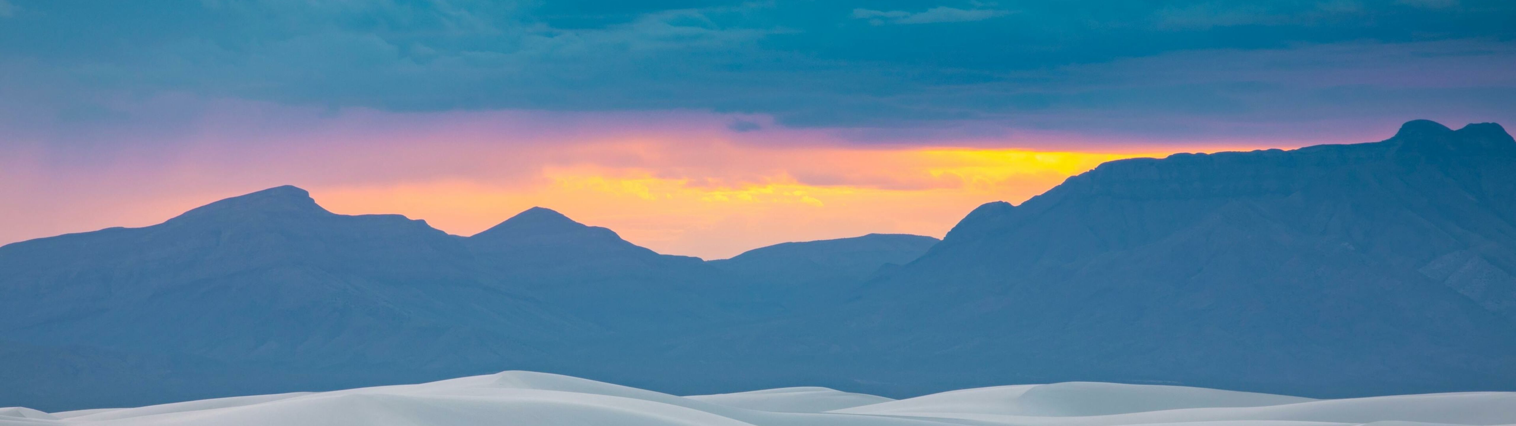 A sunset over sand dunes with mountains in the background. - 5120x1440