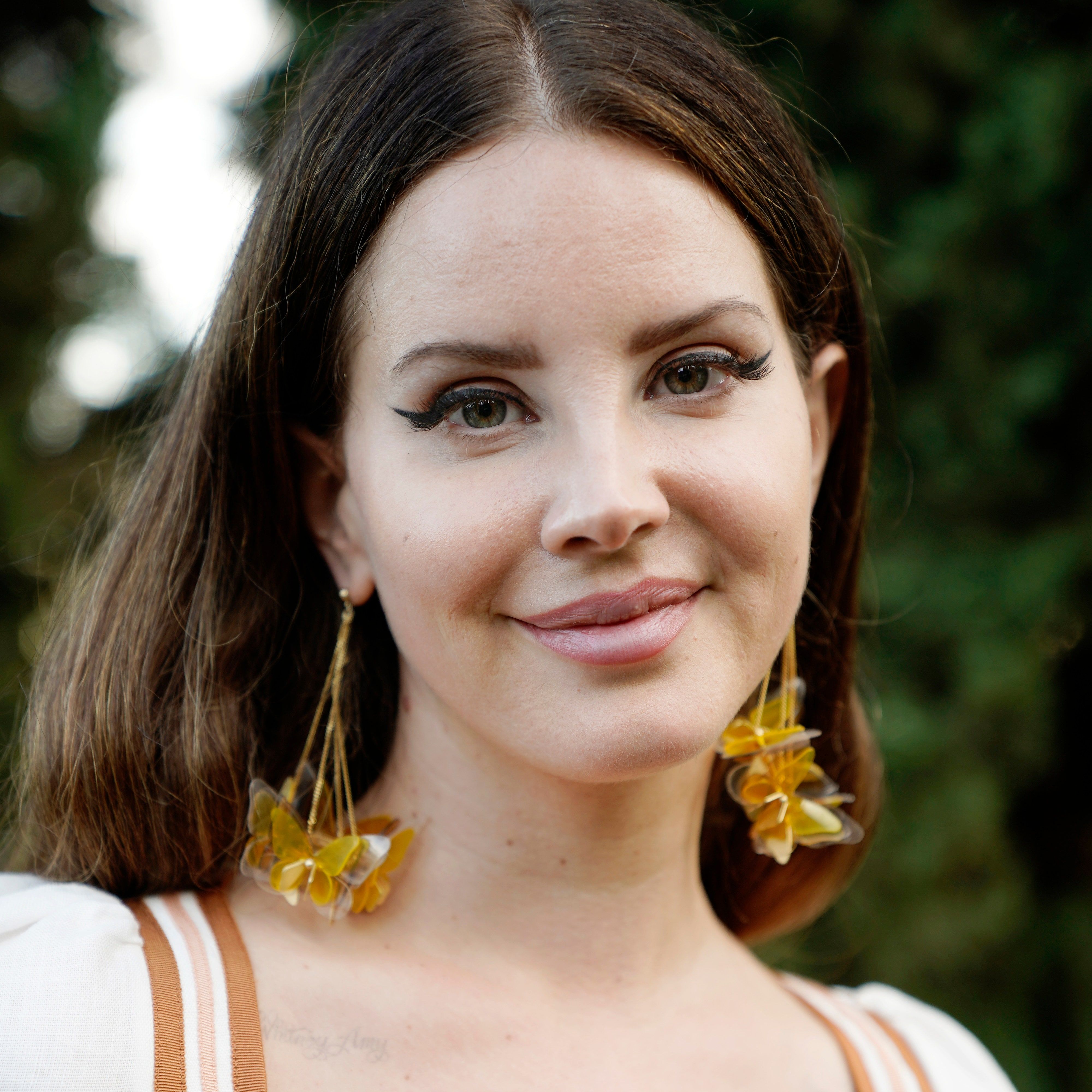 Close up of a woman with long brown hair and green eyes wearing yellow earrings - Lana Del Rey