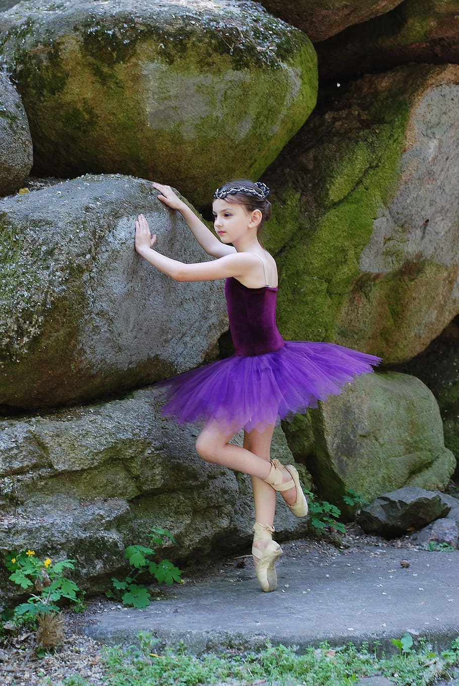 A young ballerina in a purple tutu dances in front of a rock wall. - Ballet