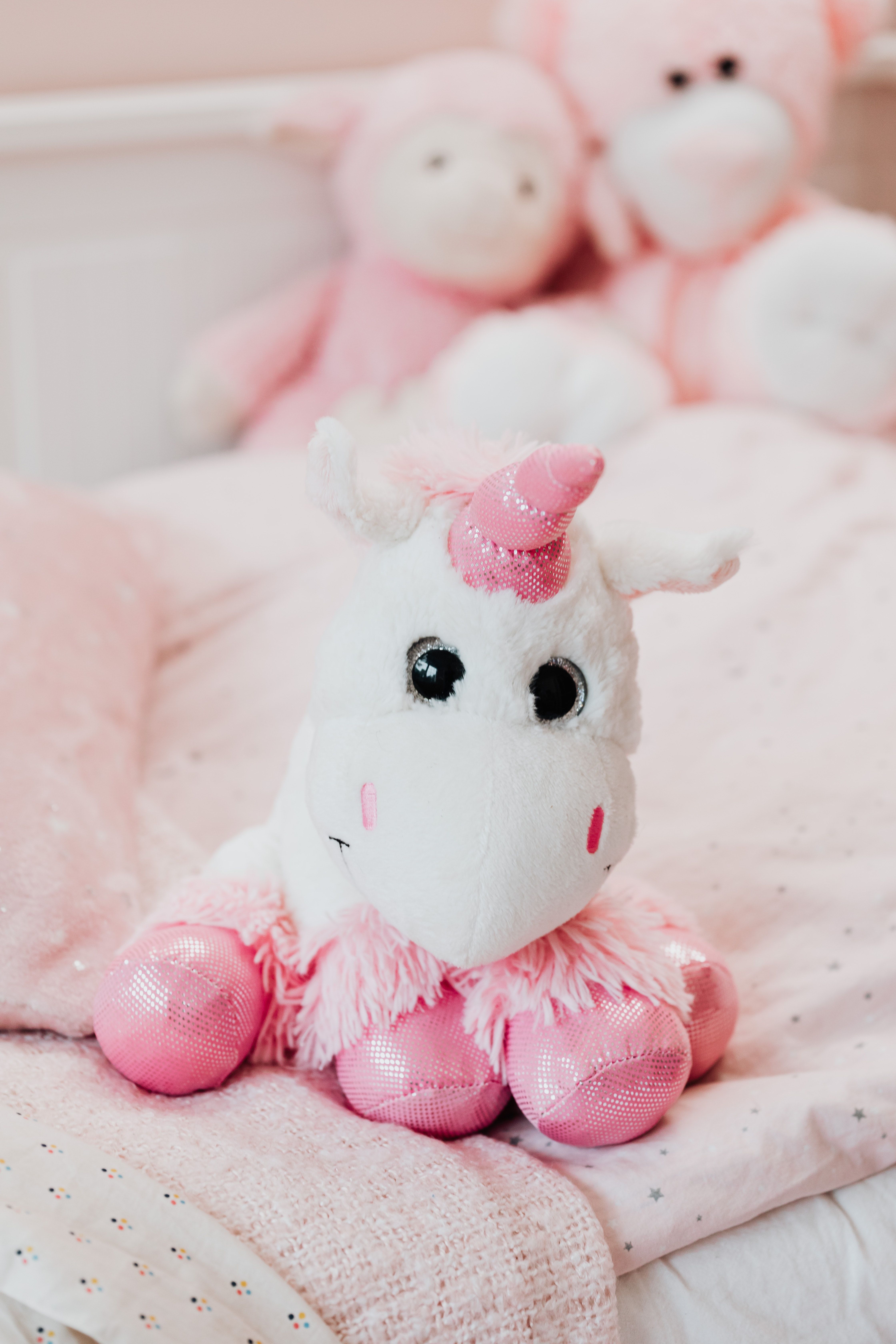 A white stuffed unicorn with a pink horn and tail sits on a bed. - Unicorn