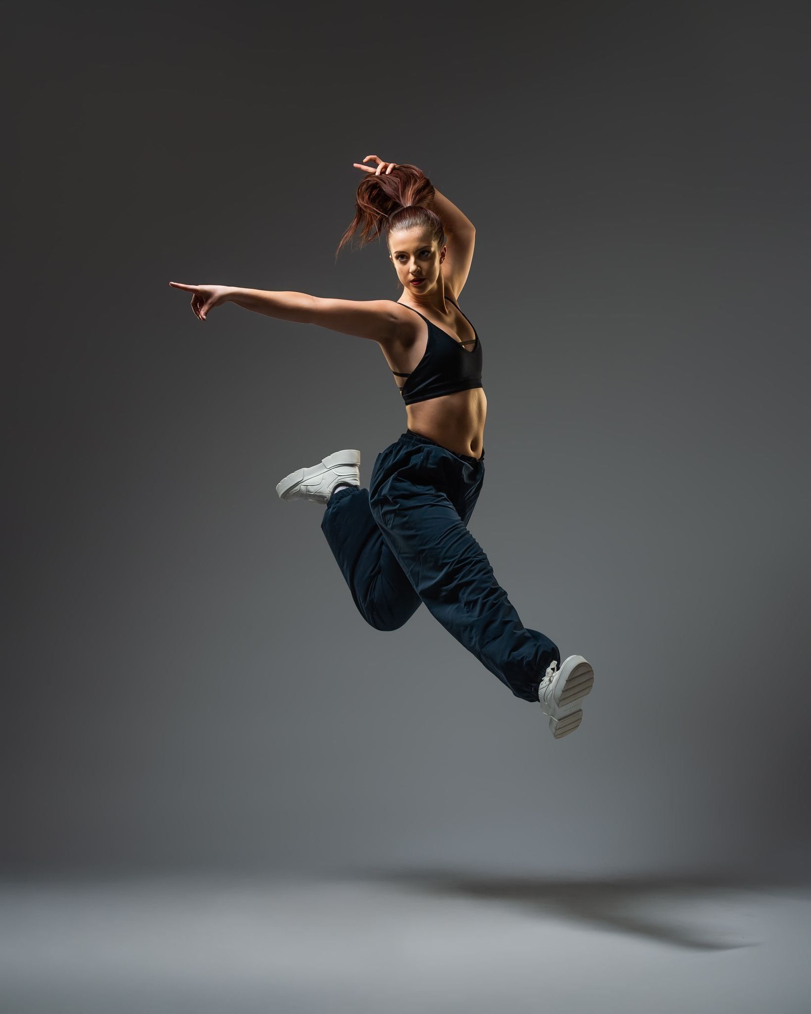 A woman in a black sports bra and sweatpants jumps in the air with her arms outstretched. - Dance