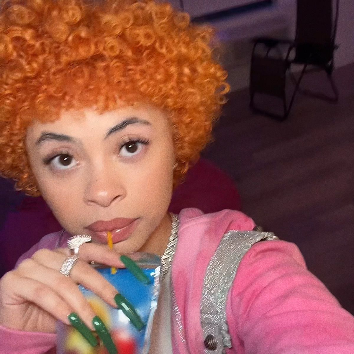 A selfie of a woman with orange hair and pink jacket holding a colorful drink. - Ice Spice