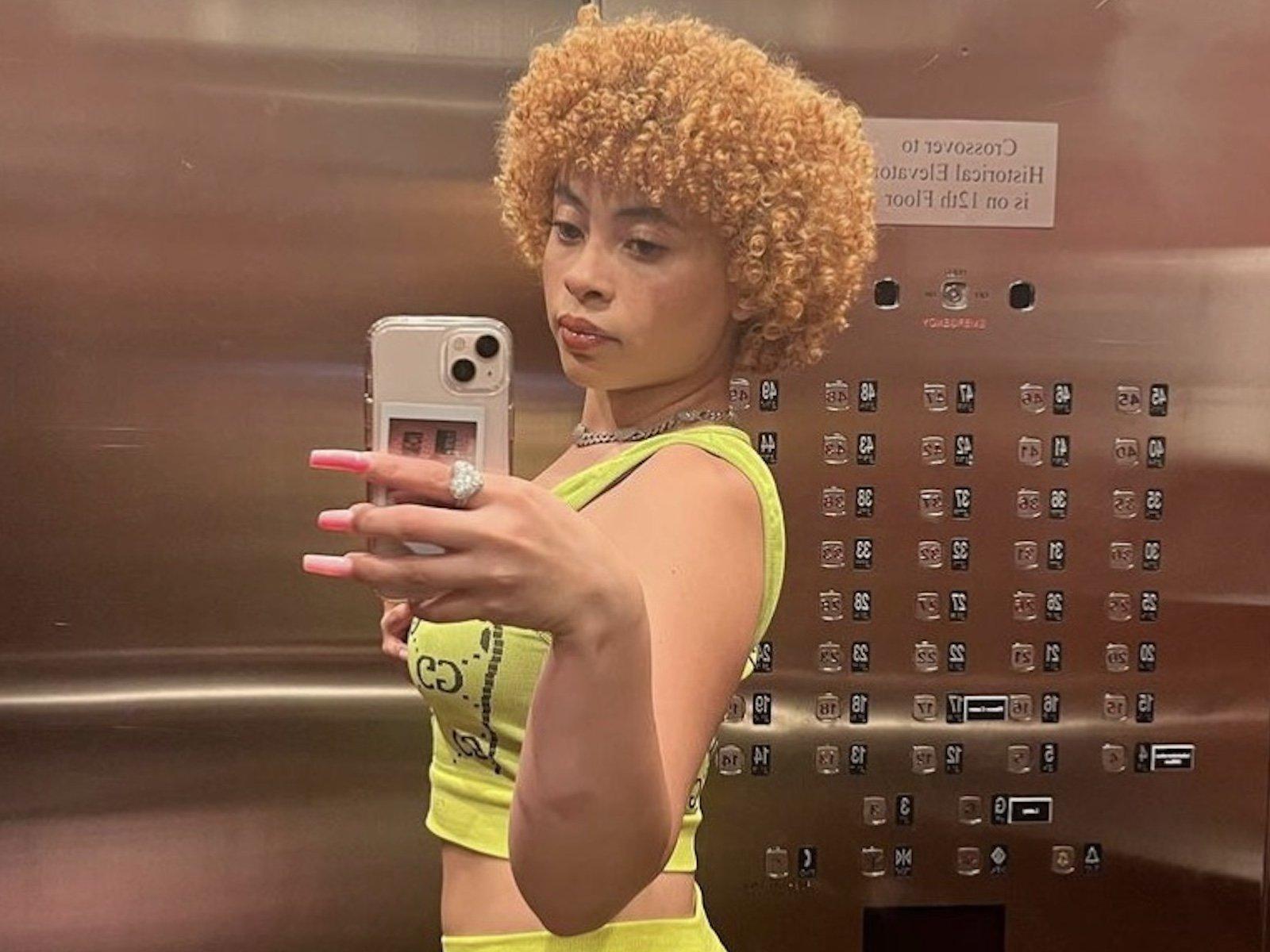 Selfie of a woman with blonde curly hair, wearing a neon green crop top and shorts. - Ice Spice