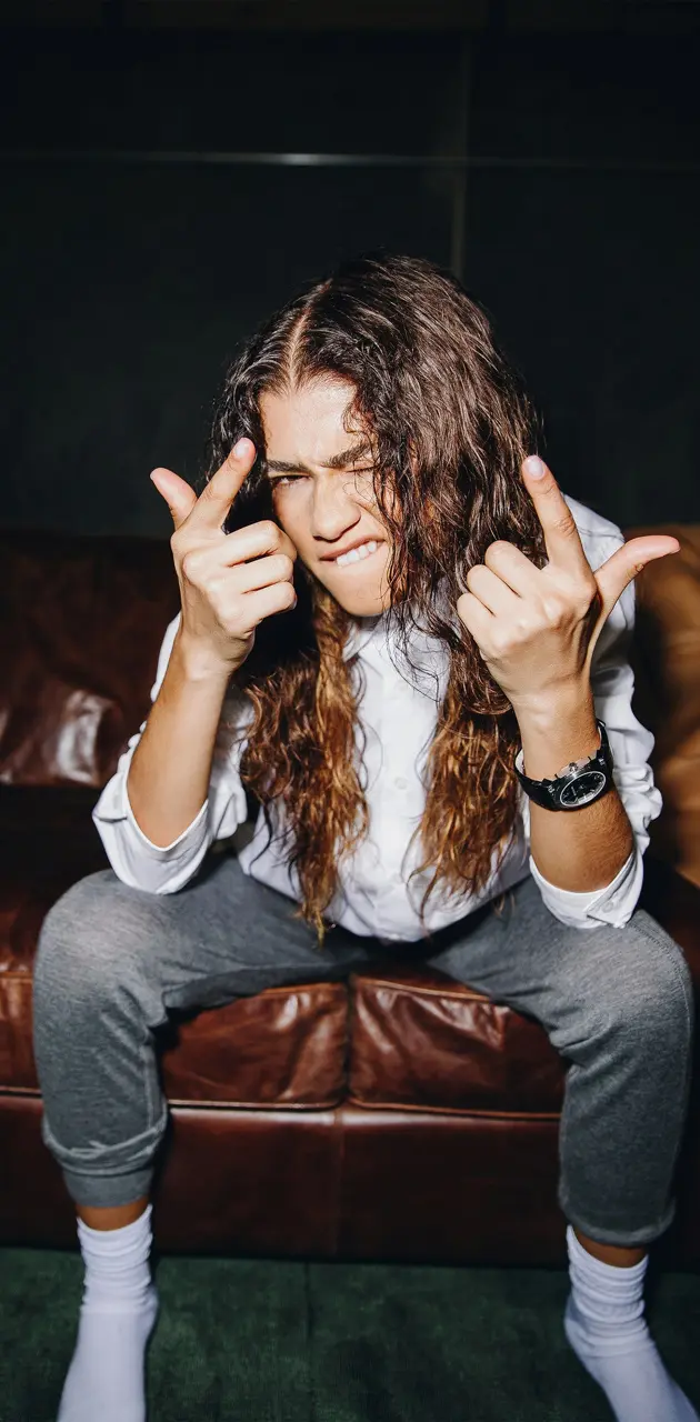 A man with long hair sitting on a couch making a rock sign with his hands - Zendaya