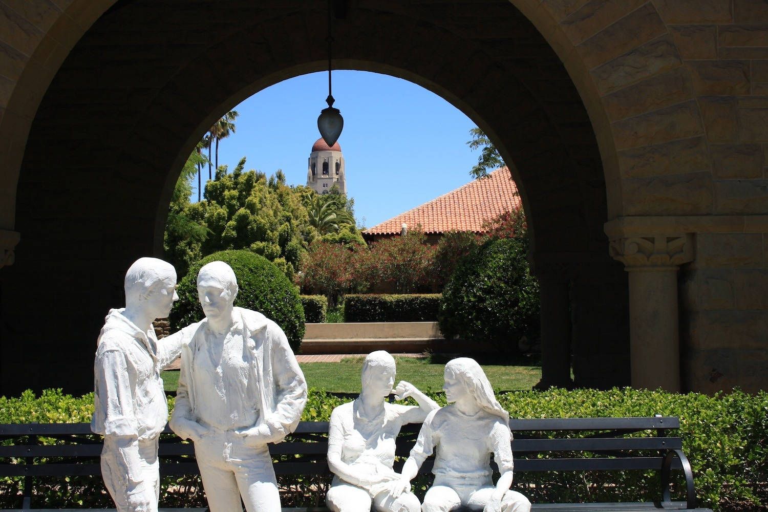 Four white sculptures of students sit on a bench in front of a red-tiled roof and a Stanford clock tower. - Stanford