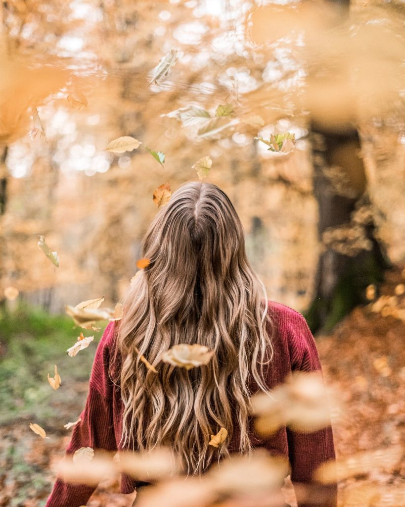 A woman with long brown hair standing in a forest with leaves falling around her - Fall