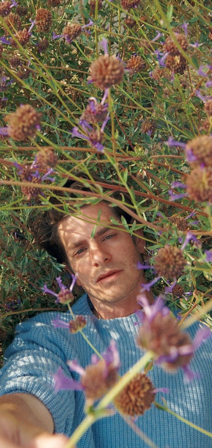 Man in blue shirt standing in front of purple flowers. - Andrew Garfield