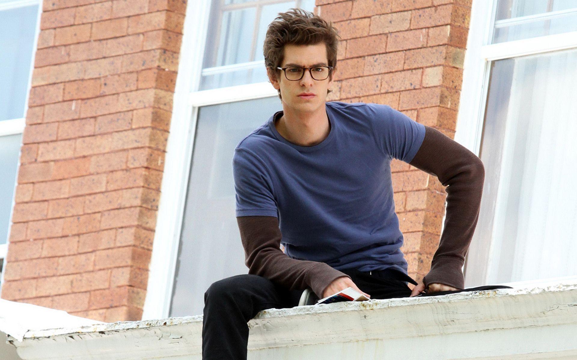 Andrew Garfield as Peter Parker in The Amazing Spider-Man. The young actor is sitting on a ledge in front of a brick building. - Andrew Garfield