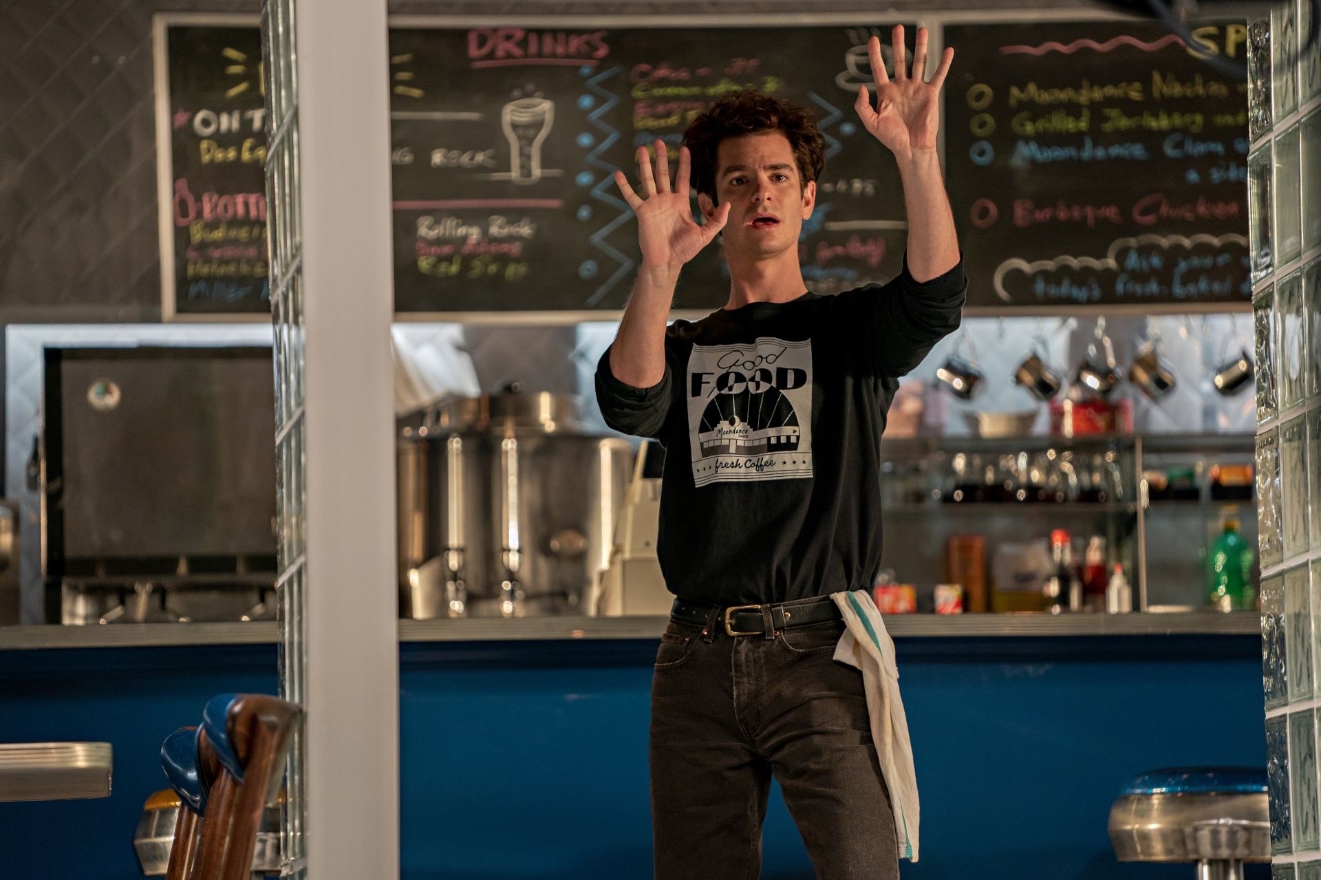 A man standing in front of a counter with his hands up in the air. - Andrew Garfield