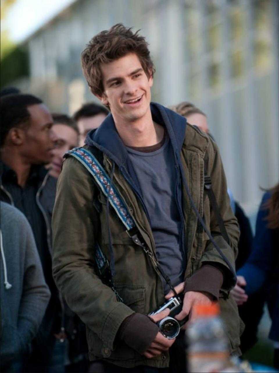 Andrew Garfield as Peter Parker/Spider-Man in The Amazing Spider-Man. He is wearing a green jacket and smiling while holding a camera. - Andrew Garfield