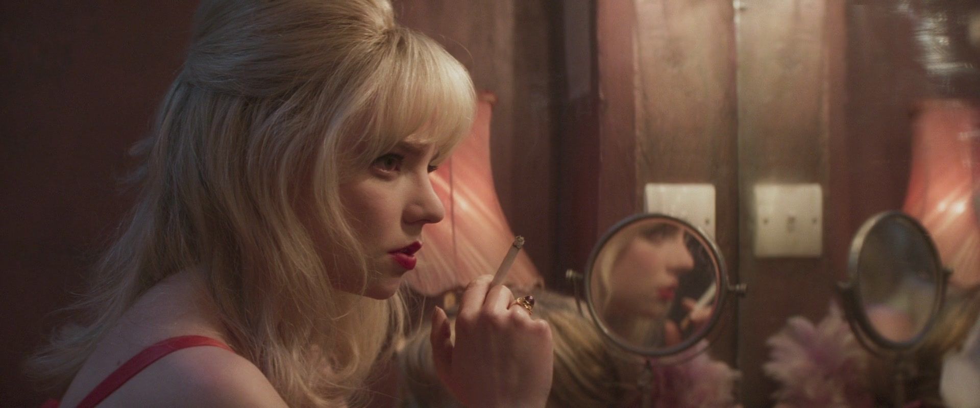 A woman with blonde hair and bangs is putting on lipstick in front of a mirror. - Anya Taylor-Joy