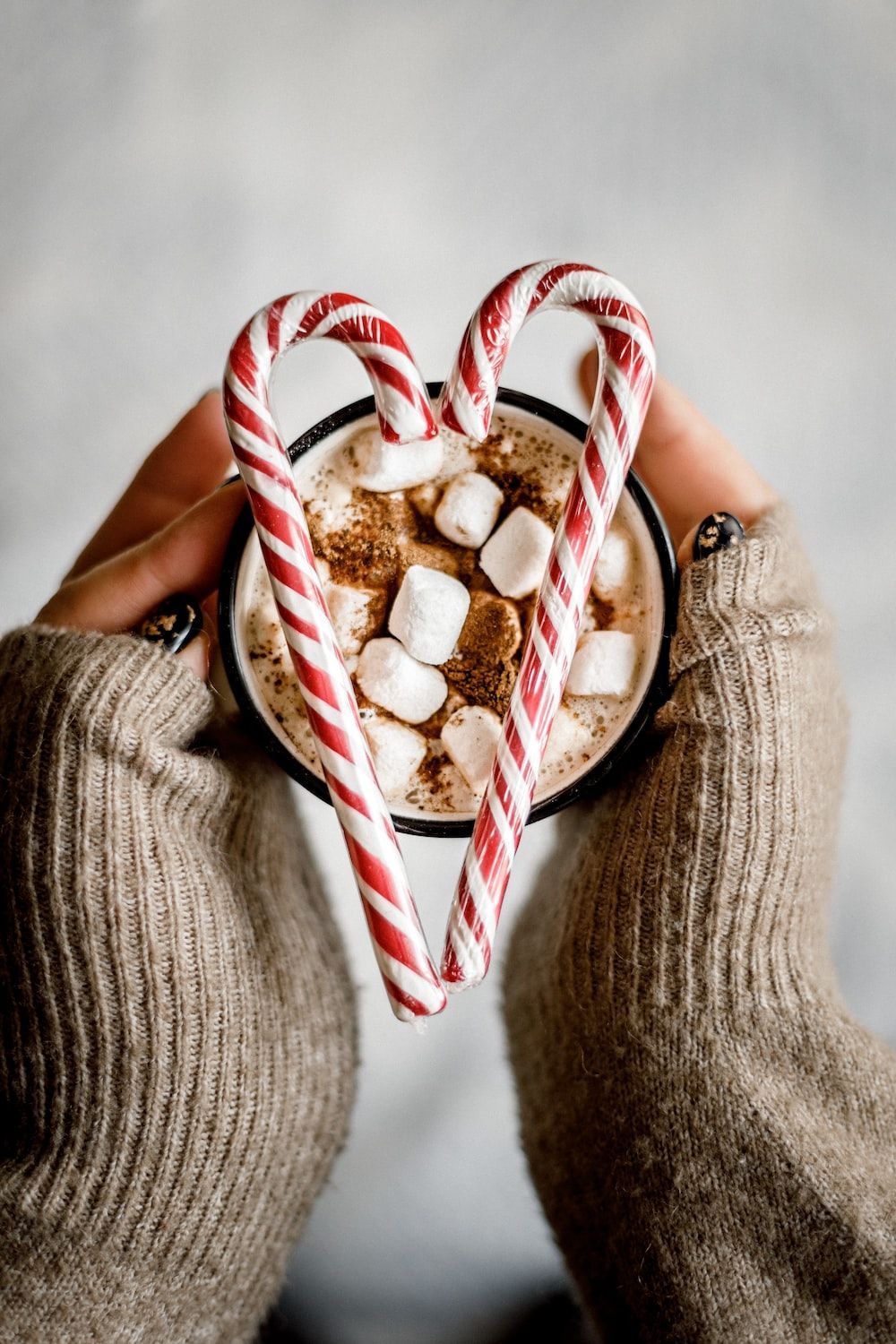 A woman's hands holding a mug of hot chocolate with candy canes in the shape of a heart. - Marshmallows