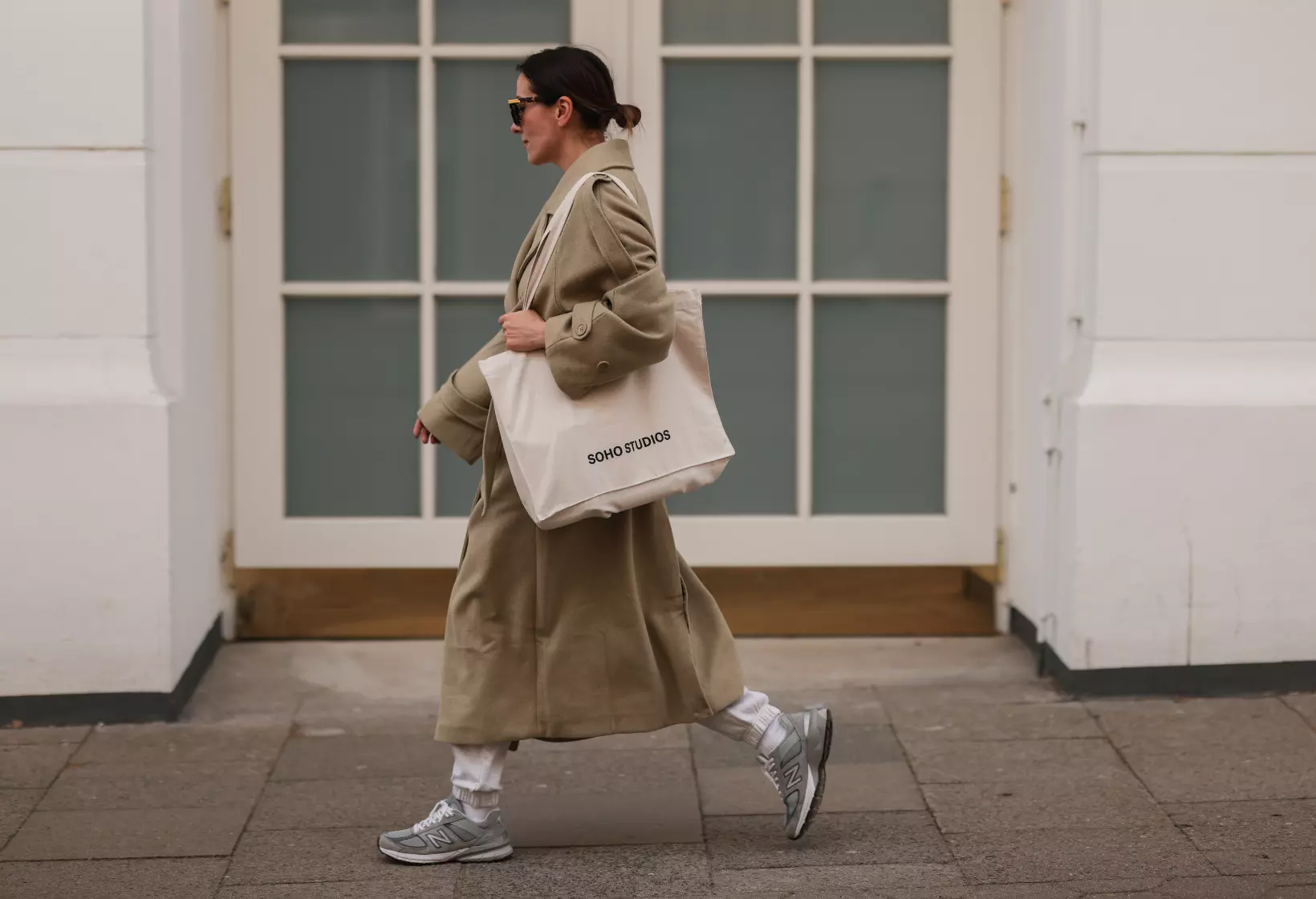 A woman in a beige coat and white bag walking down the street. - New Balance