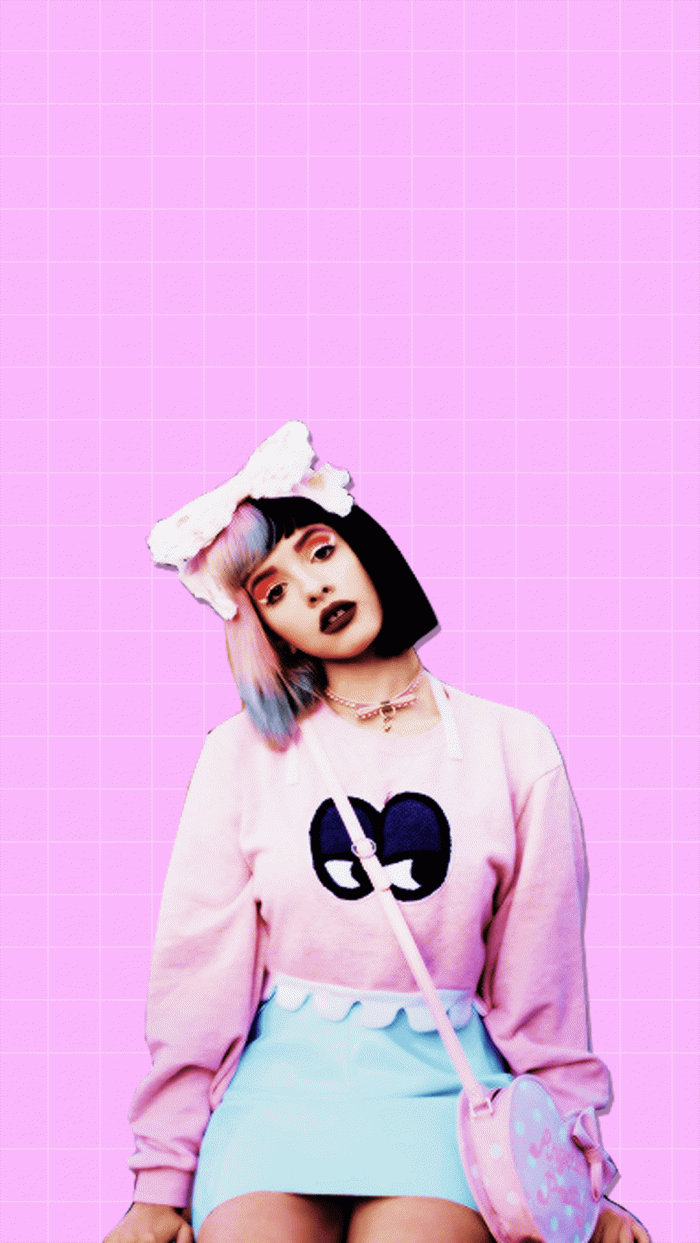 A woman with a pink and blue outfit and a bow in her hair sits on a stool in front of a pink grid background. - Melanie Martinez