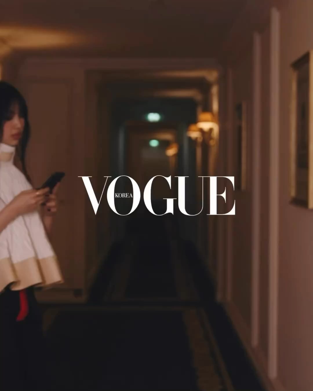 A woman in a white dress stands in a hallway holding a phone. - Vogue