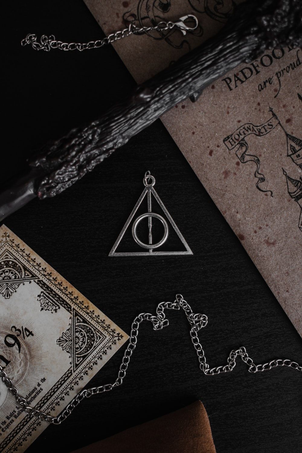 A Deathly Hallows necklace on a black background with a wand and a book. - Hogwarts
