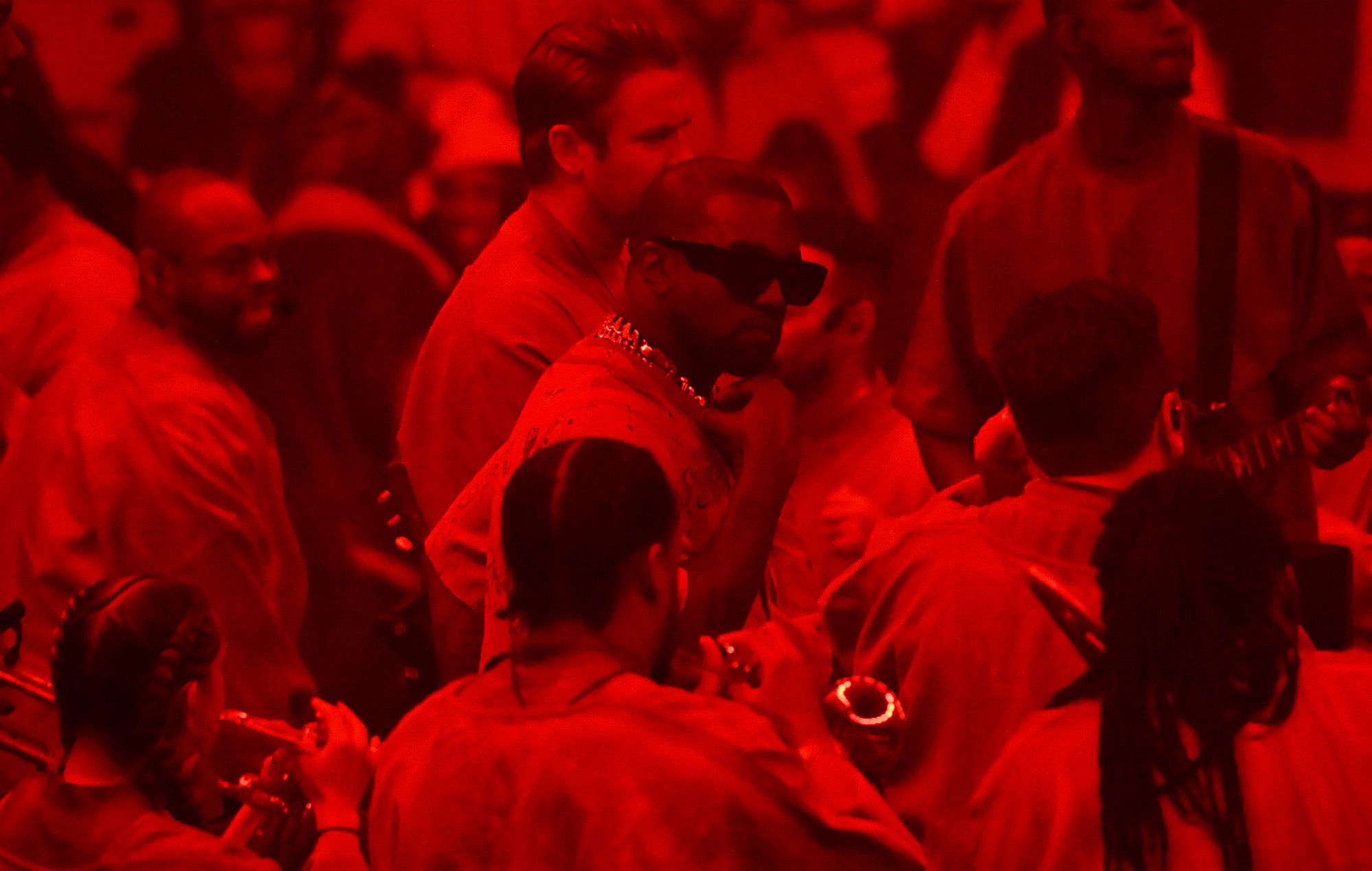 Kanye West, center, with a red filter over the image, surrounded by people at a party. - Kanye West